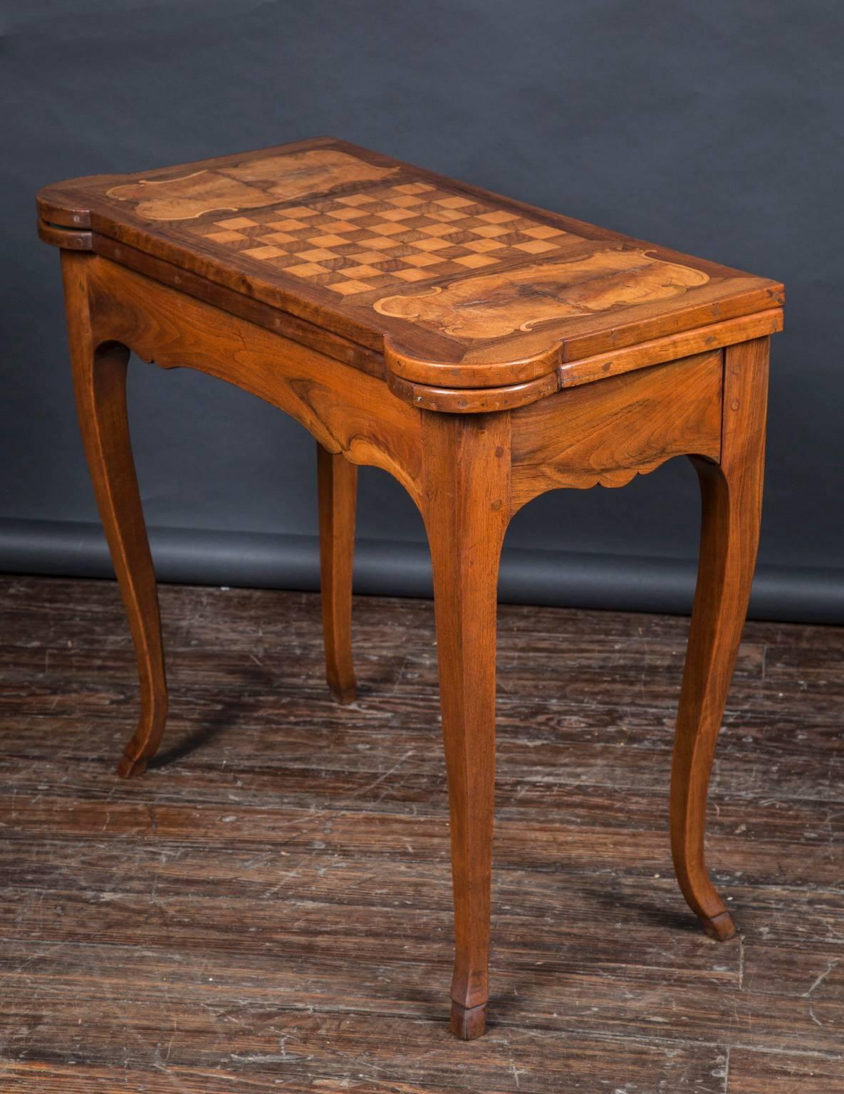 Cette table de jeu du XVIIIe siècle est fabriquée en bois fruitier et son plateau est orné d'un damier incrusté. La table dispose d'un espace de rangement pour les pièces de jeu sous le plateau, et le plateau se replie pour révéler une surface de