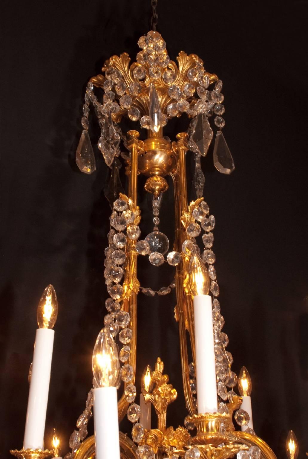This antique chandelier is made of gold covered bronze, and features 2 staggered tiers of lighting, with each light resting atop a highly decorated bobeche. The lights are mostly grouped in sets of fours, with three lights in the lower tier resting