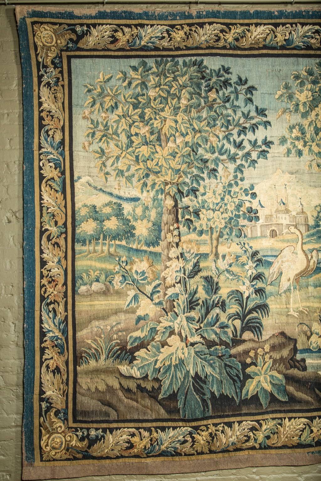 This grand French Aubusson Verdure Tapestry dates back to the 18th century and features an exotic landscape with mythical animals. A long legged bird forages amidst exotic blossoming foliage at the center. Behind it rests a grand hillside with