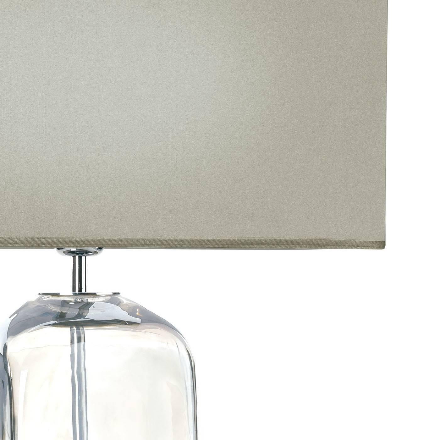 The chromed metal of the shaft of this lamp is visible through the transparency created by the glass container that envelops it. This striking base makes the fabric-covered rectangular shade above seemingly float in space and gives lightness to this