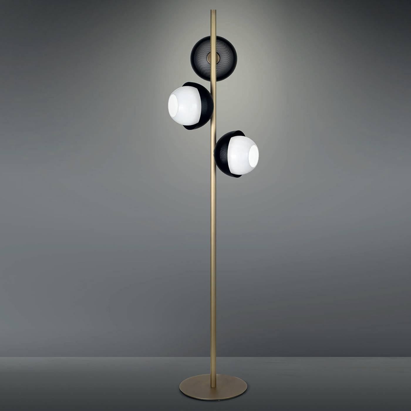 This striking floor lamp features a minimalist shaft in brass, which can have a light or dark burnished brass finish, resting on a round base of the same material and finish. The three shades that stem from the top face different directions creating