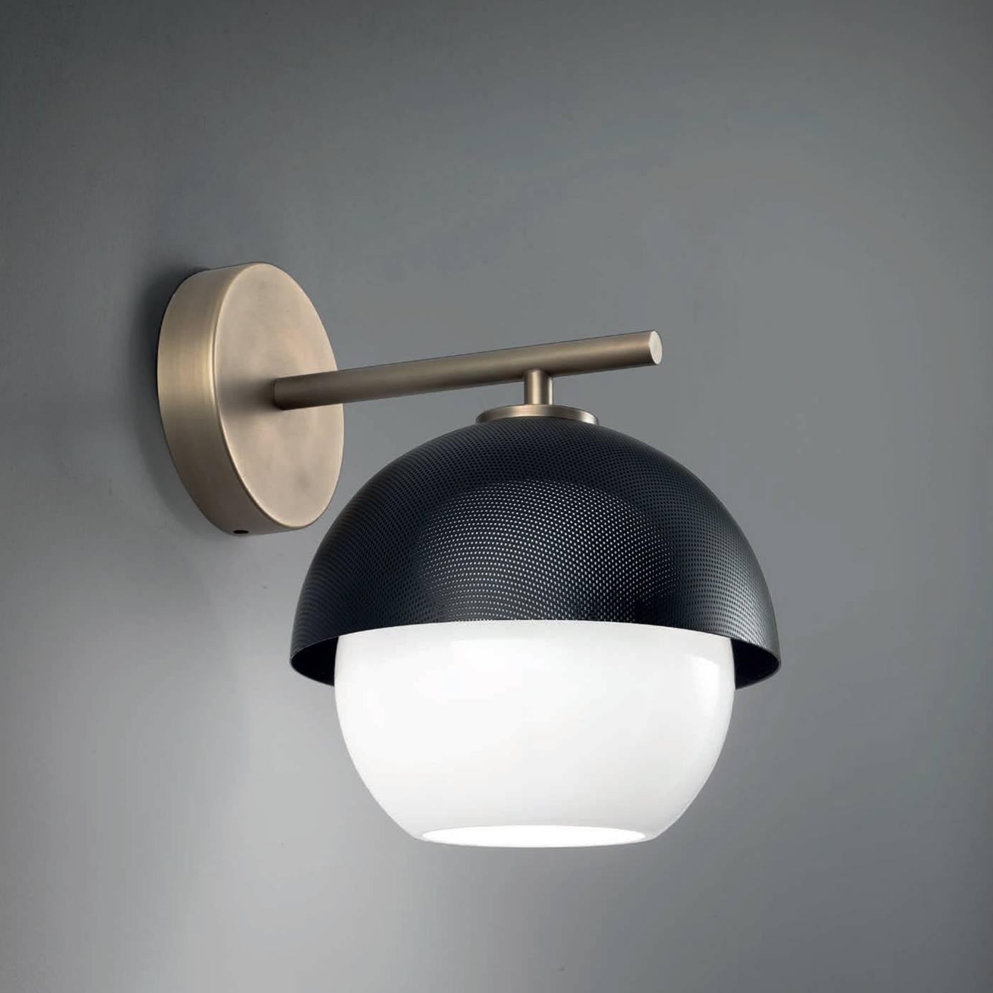 This sophisticated sconce has a brass structure in a light or dark burnished finish that supports a semi-spherical shade facing downwards and made in metal that can be either given a matte black or a matte golden finish, for a warmer final effect.