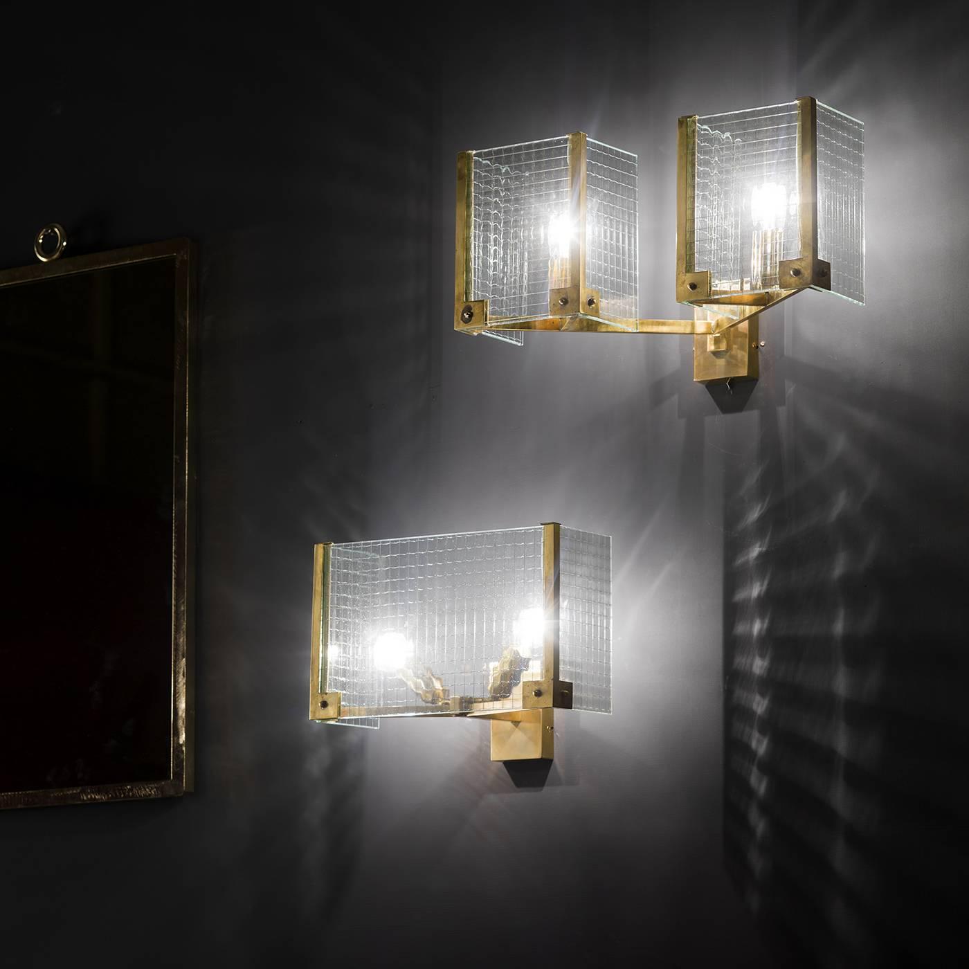 This exquisite sconce is anchored to the wall using a rectangular brass base. Extending outwards from the base are two arms, also in natural brass, which hold each of the two illuminating elements of the piece. Surrounding each light is a