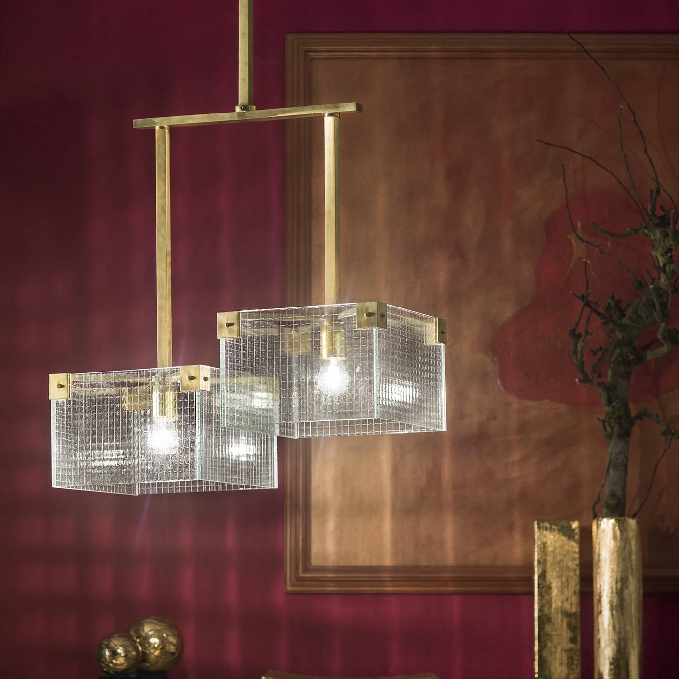 This chandelier features a unique asymmetrical structure crafted in natural brass. One horizontal brass rod is connected to two vertical ones of different lengths, each one holding an illuminating element. The two rectangular shades are in