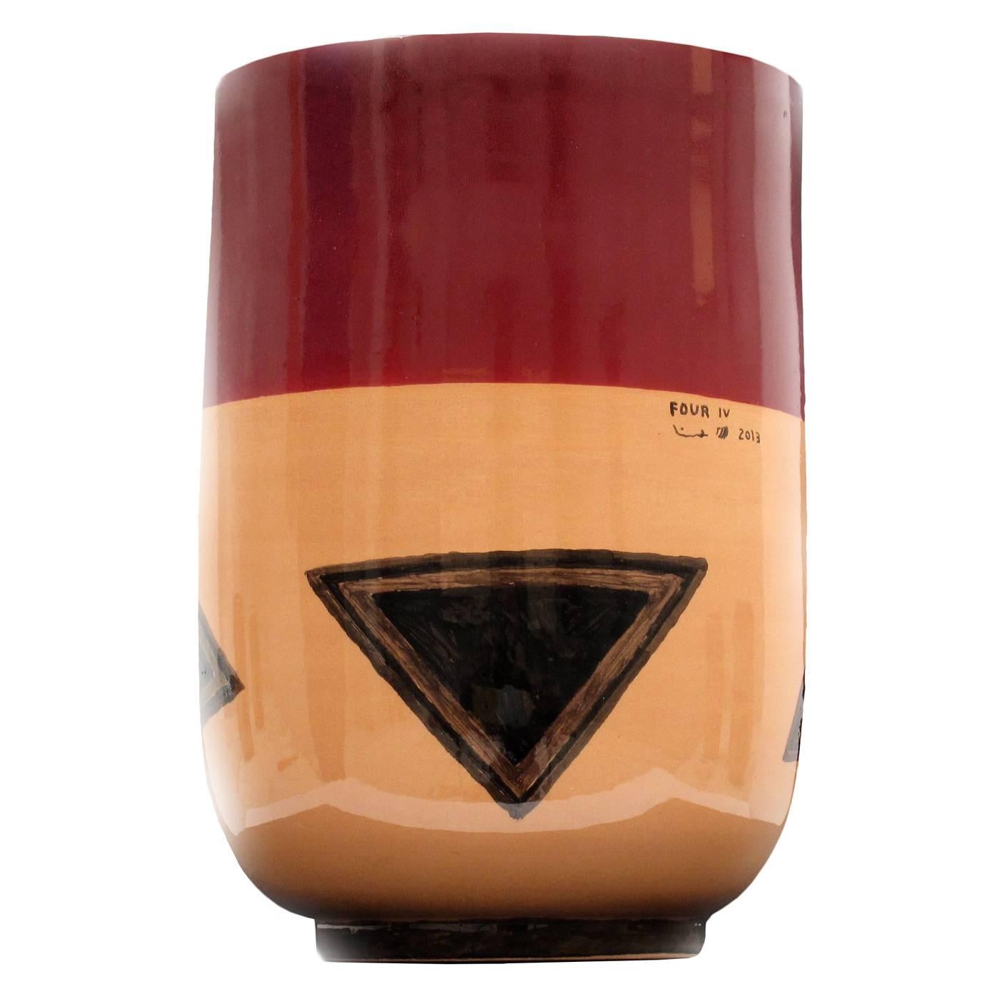 This exquisite vase is made entirely of ceramics. Its small plinth base supports a cylinder that curves at the base to resemble a large cup. The smooth surface of the piece is in a color block combination of deep maroon and rich orange, on which are