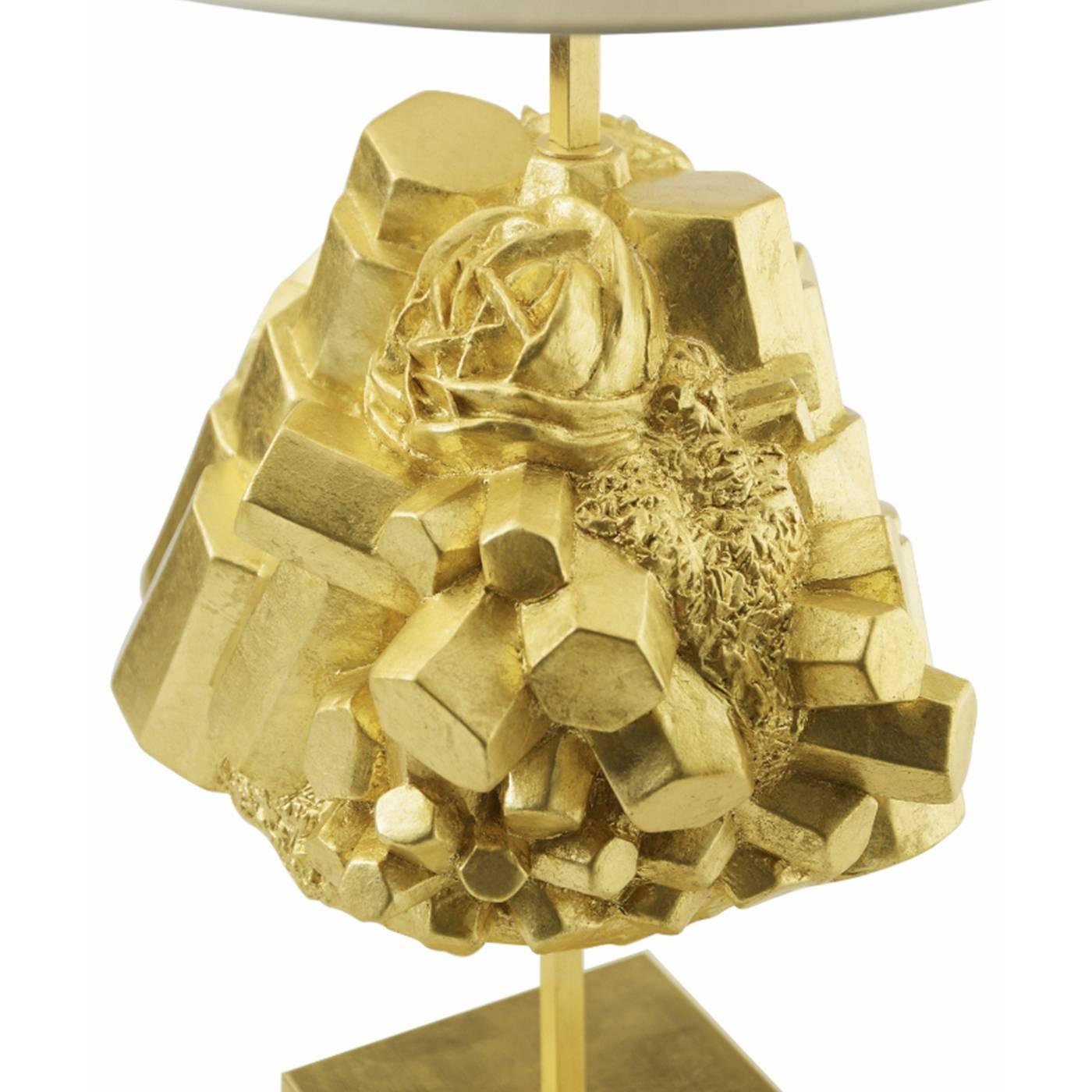 This striking table lamp features a metal structure resting on a gold finished square base that supports a white fabric shade. The standout element of the piece is a sculpture used as decorative accent and made entirely of ceramic with a gold finish