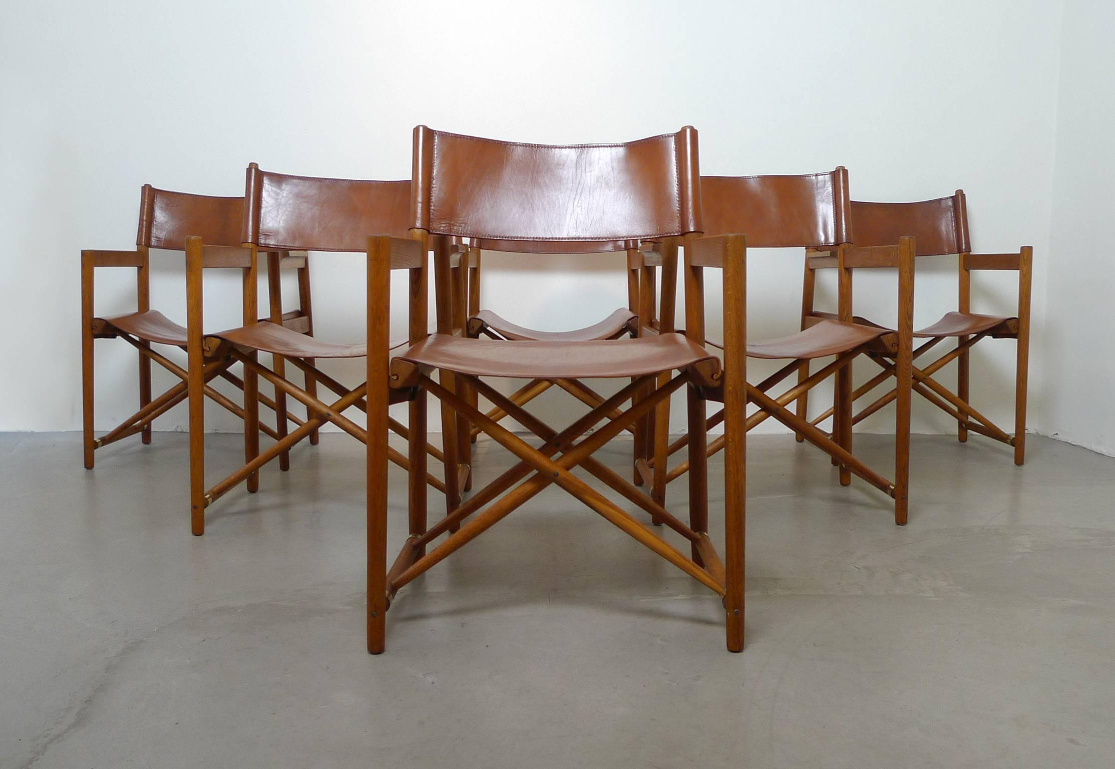 Rare foldable Safari chairs with an oak frame, cognac brown neck leather and brass sleeves. The dining chairs are in very good vintage condition, the leather surface has a charming patina.