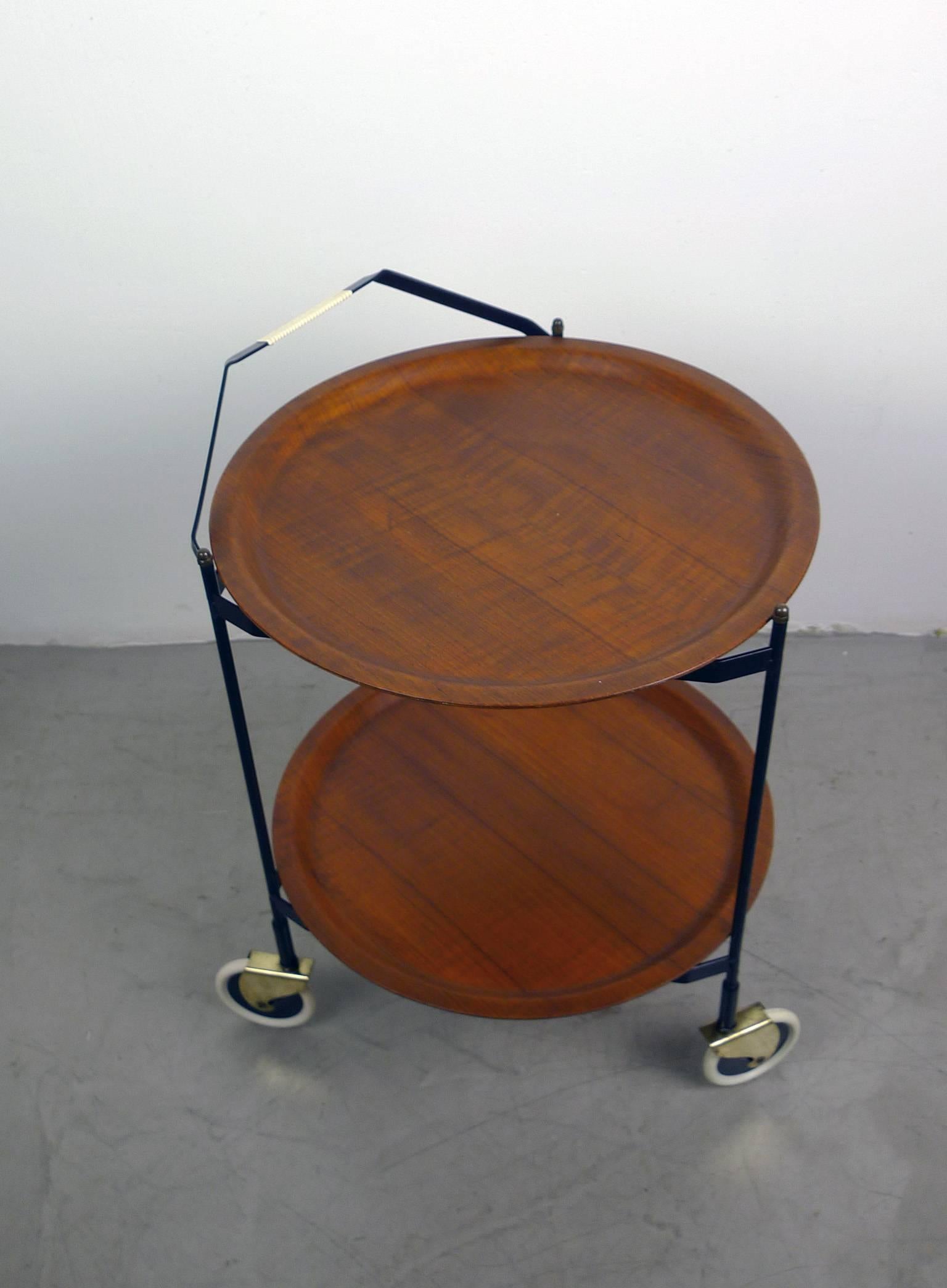 This mobile tea trolley with two removable round teak trays has a black lacquered metal frame that is foldable for space-saving storage. The three rolling feet are fixed to the rack by brass elements. It was produced in the 1950s in Denmark and is