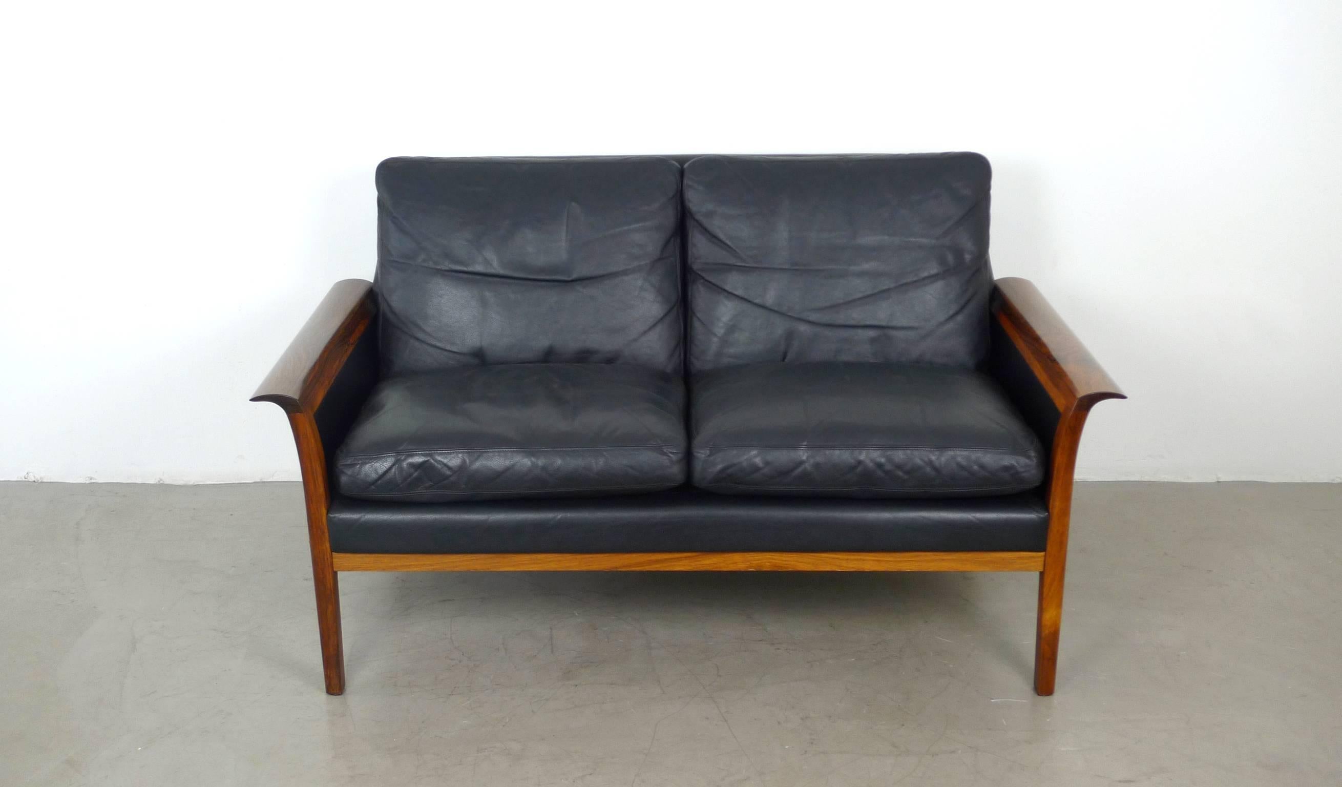 Elegant black leather sofa with rolling rosewood frame for two persons. All seat and backrest cushions are filled with down feathers for a soft and comfortable sitting. The frame of the left and the right side of the sofa is made of solid