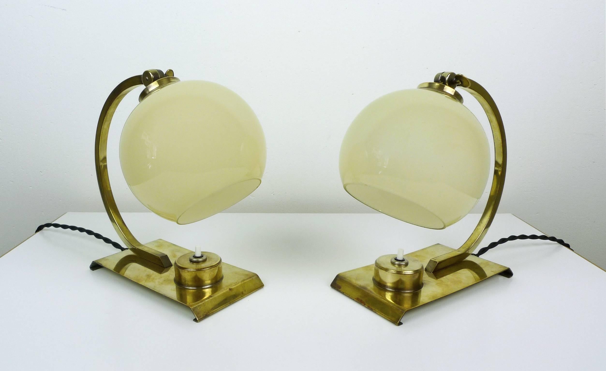 Two Art Deco table lamps from the 1930s with beige glass screens. Stand and bracket are made of brass.
The inclination of the ball screen can be adjusted by a small wing screw at the upper end of the bracket. Each lamp has a pressure switch on the