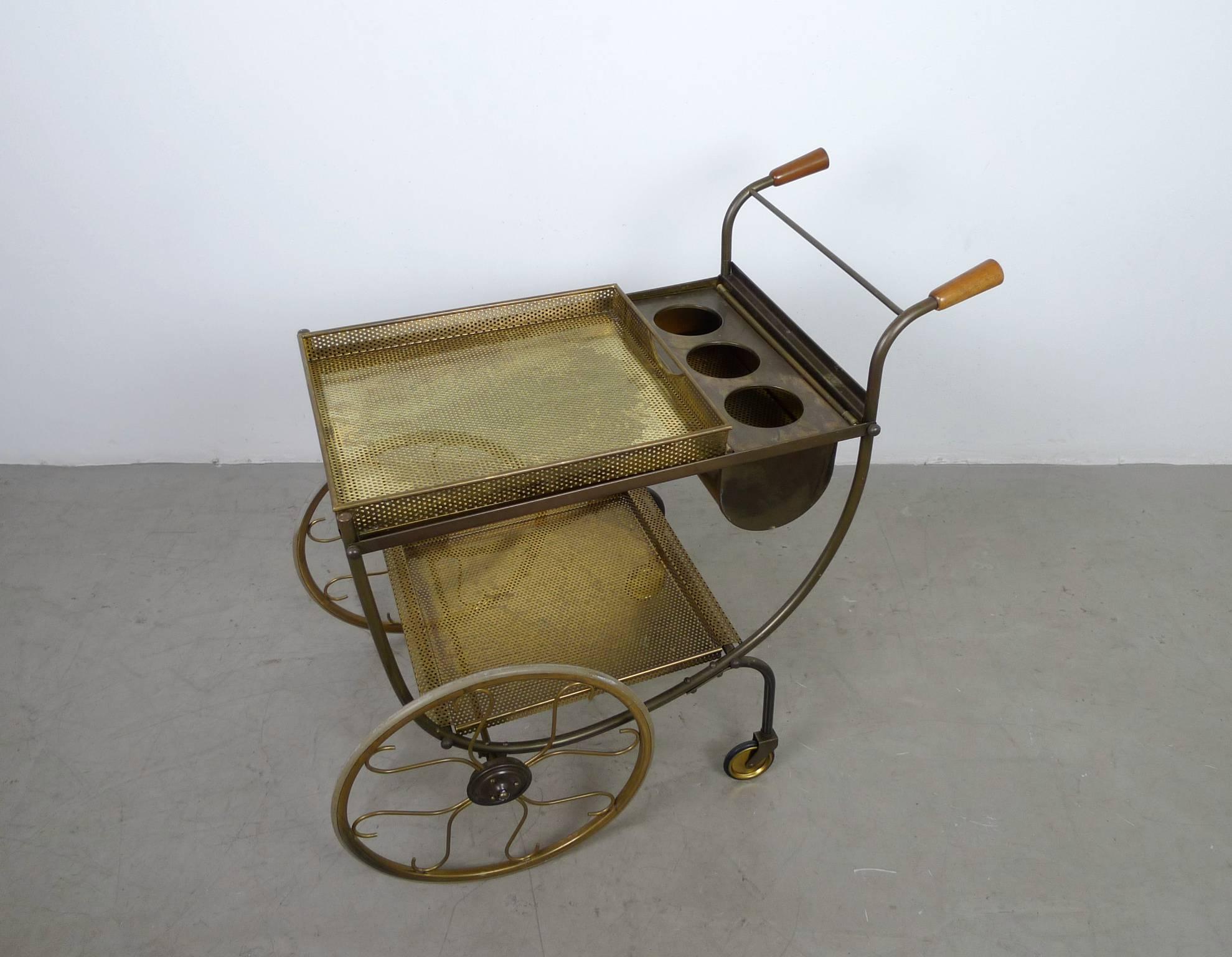 This serving trolley has large brass wheels and two wooden handles. The upper and lower shelves and the curved bottle holder are removable. The top shelf is equipped with a tray and carrying handles for serving. The trolley was designed by Josef