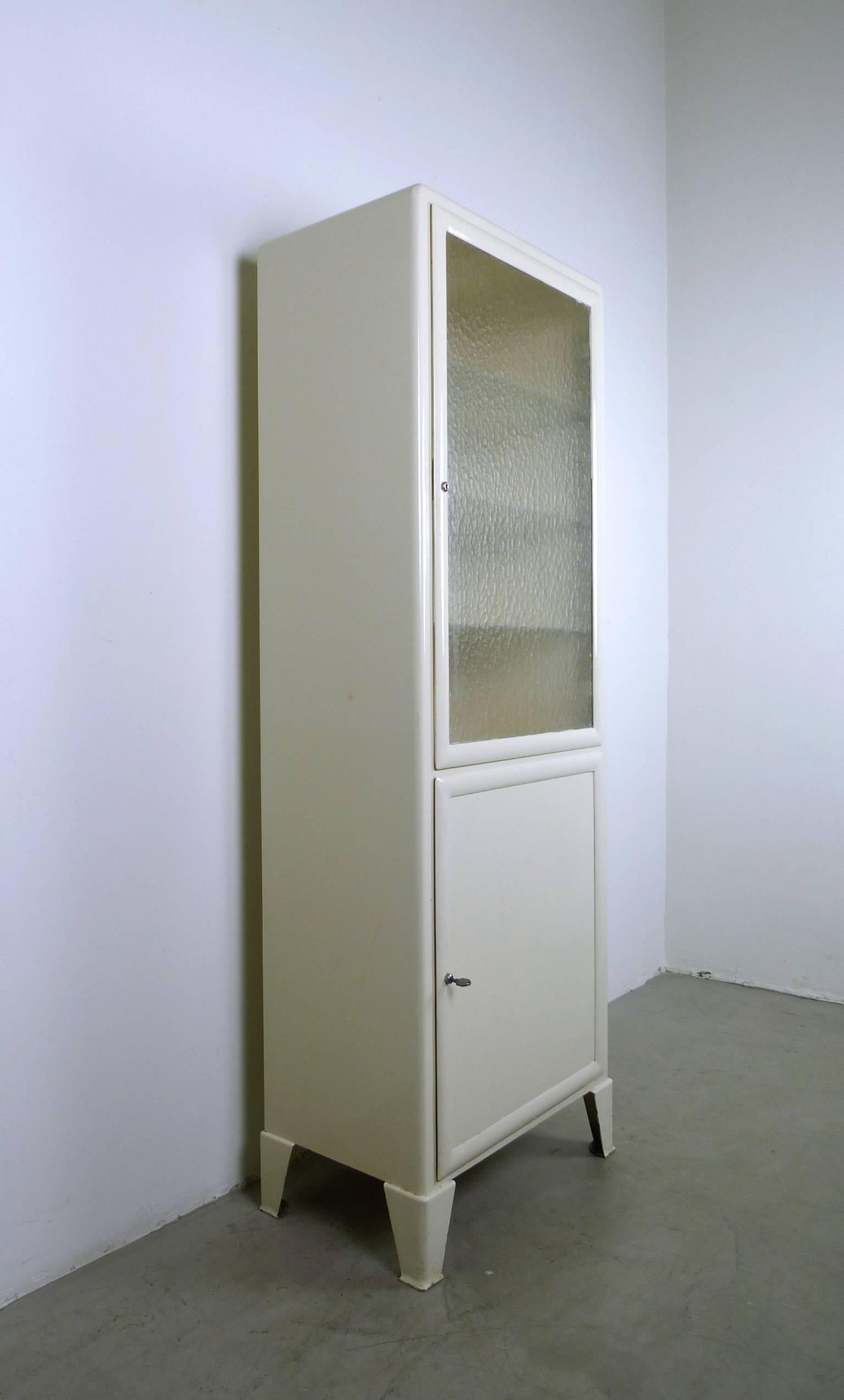 Two-door, ivory-colored steel cabinet with cathedral glass pane embedded in the upper door. Both doors equipped with locks, the original key is present. Behind the upper door are three height-adjustable shelves in corrugated glass. Behind the lower