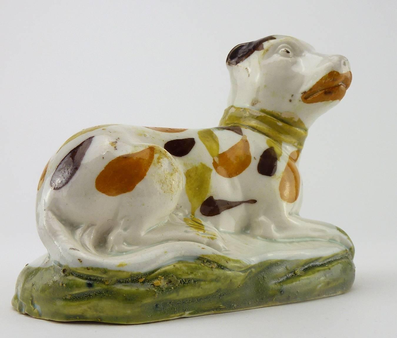 English Yorkshire pearlware prattware figure of recumbent hound dog, modelled on a low-mounded ovular base, painted green. The white dog with ocher, yellow, brown spltches, and prominent enameling to its lips, dots on eyes, and brownband over the