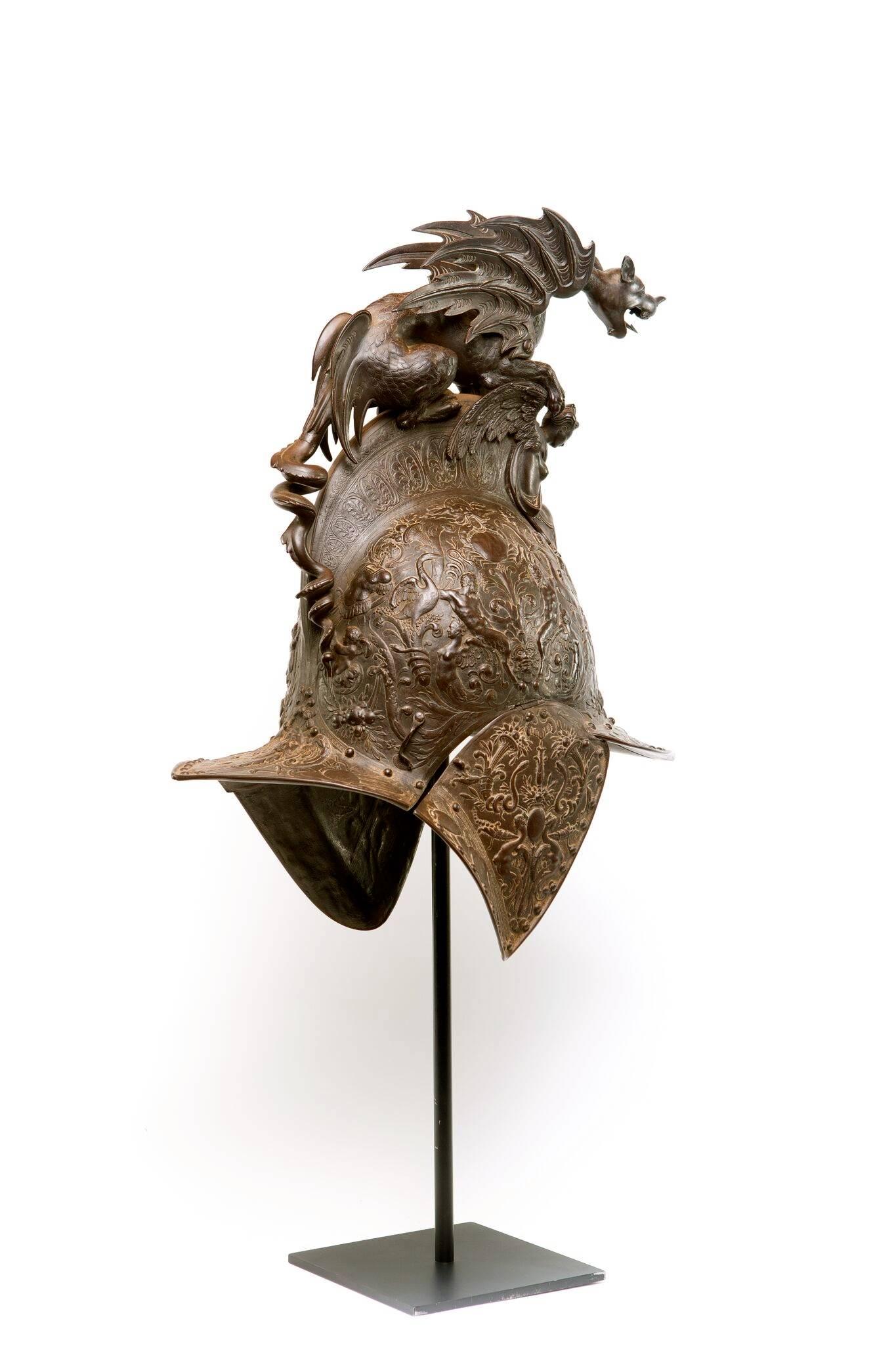 A Renaissance style relief decorated bronze metal parade helmet with pointed visor, hinged cheek flaps and flared neck cover, decorated with classical satyrs, winged terms and cranes. Surmounted by griffin crest above caryatid, on a metal pole