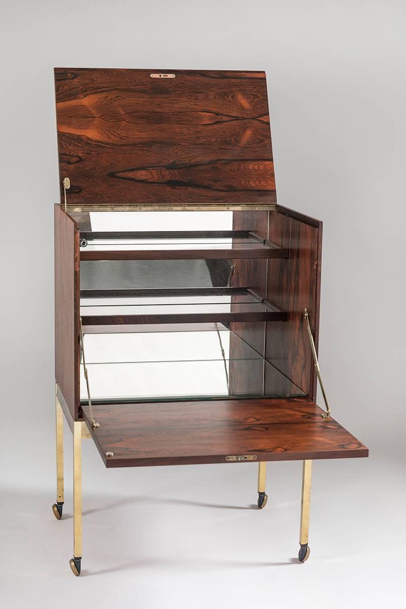 Gorgeous small bar cabinet in rosewood with legs and wheels made of brass, probably manufactured in Sweden. The top and front fold out, creating a mini bar with glass drawers and mirrored glass on the inside. 
Condition: The wood is in excellent