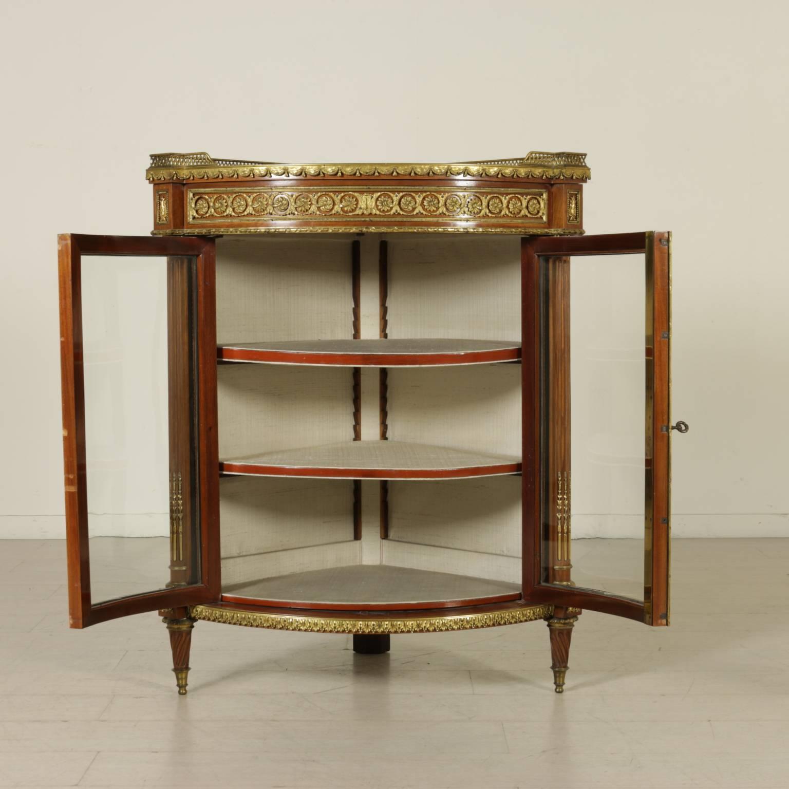 A semicircular solid mahogany corner cabinet with two doors with glasses, truncated conical feet with helical groove, half columns on the uprights, marble top enclosed by a curtain-shaped gilded bronze small baluster. The cabinet is enriched by