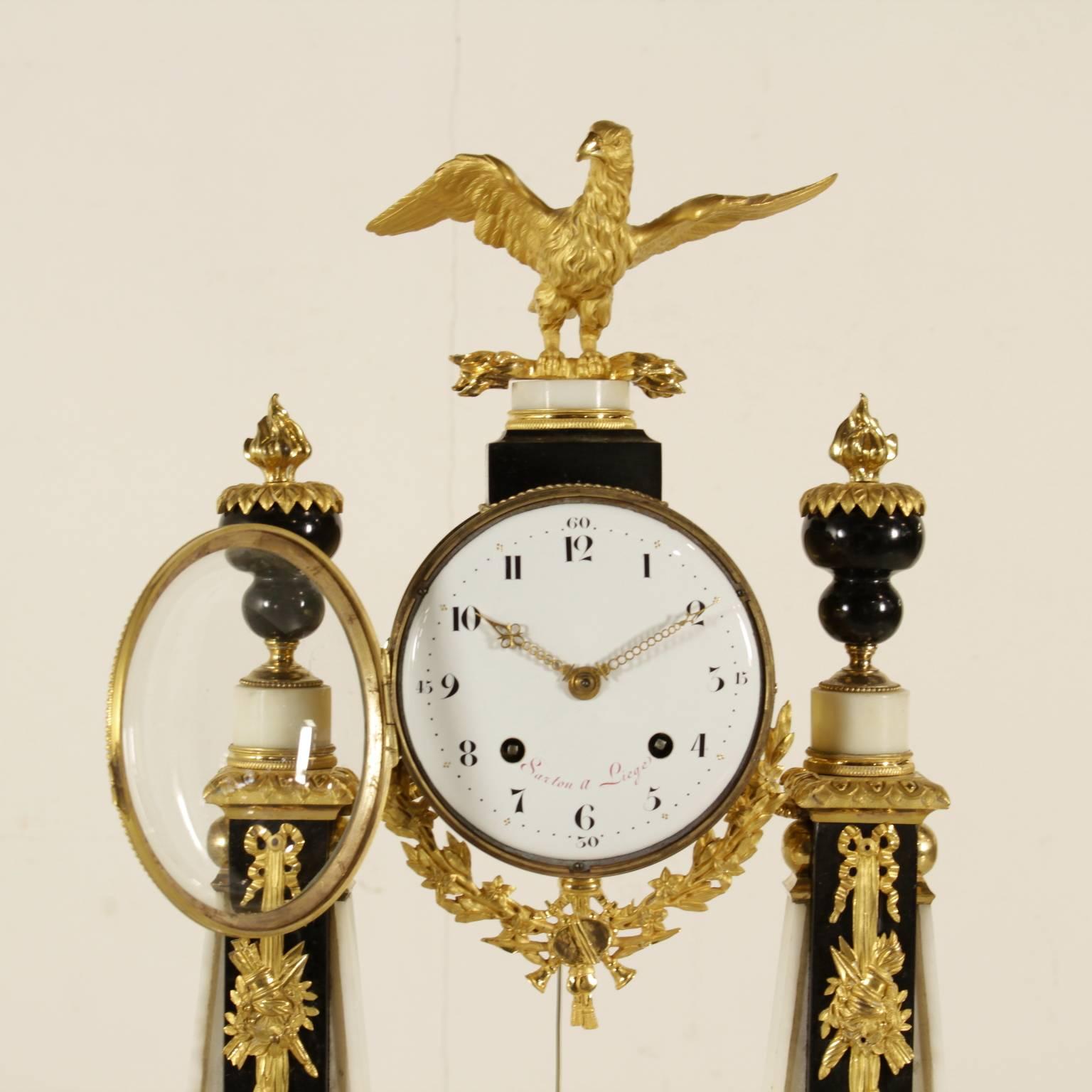 A neoclassical round clock with two white and black marble obelisks and a Carrara marble basement. The clock face is lacquered, protected by glass and marked 