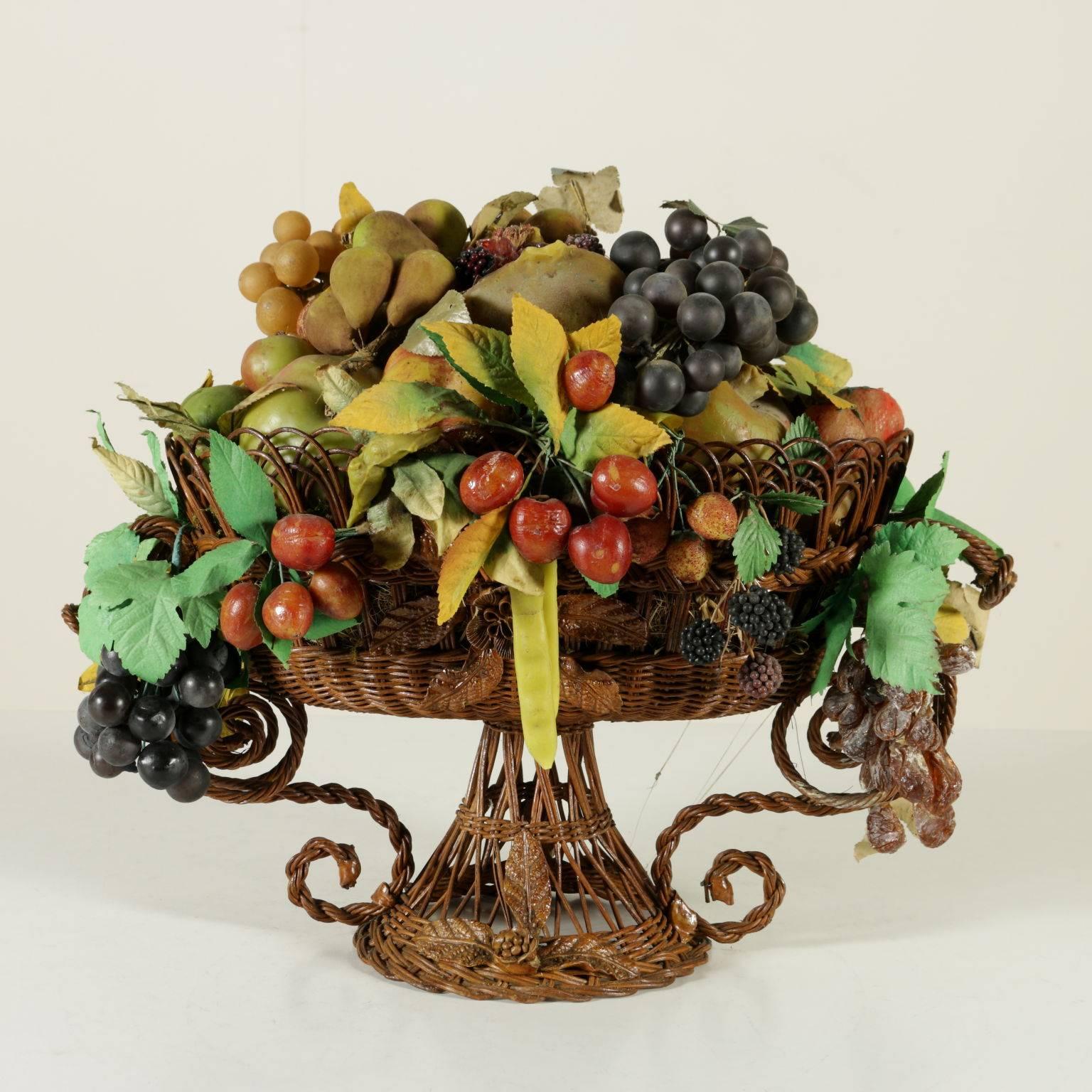 An elegant wicker basket features a colorful wax fruit composition with branches and leaves made of iron wire, paper and colored cloth. There are pomegranates, grape bunches, berries, plums, aubergines, mushrooms, etc. The realistic realization