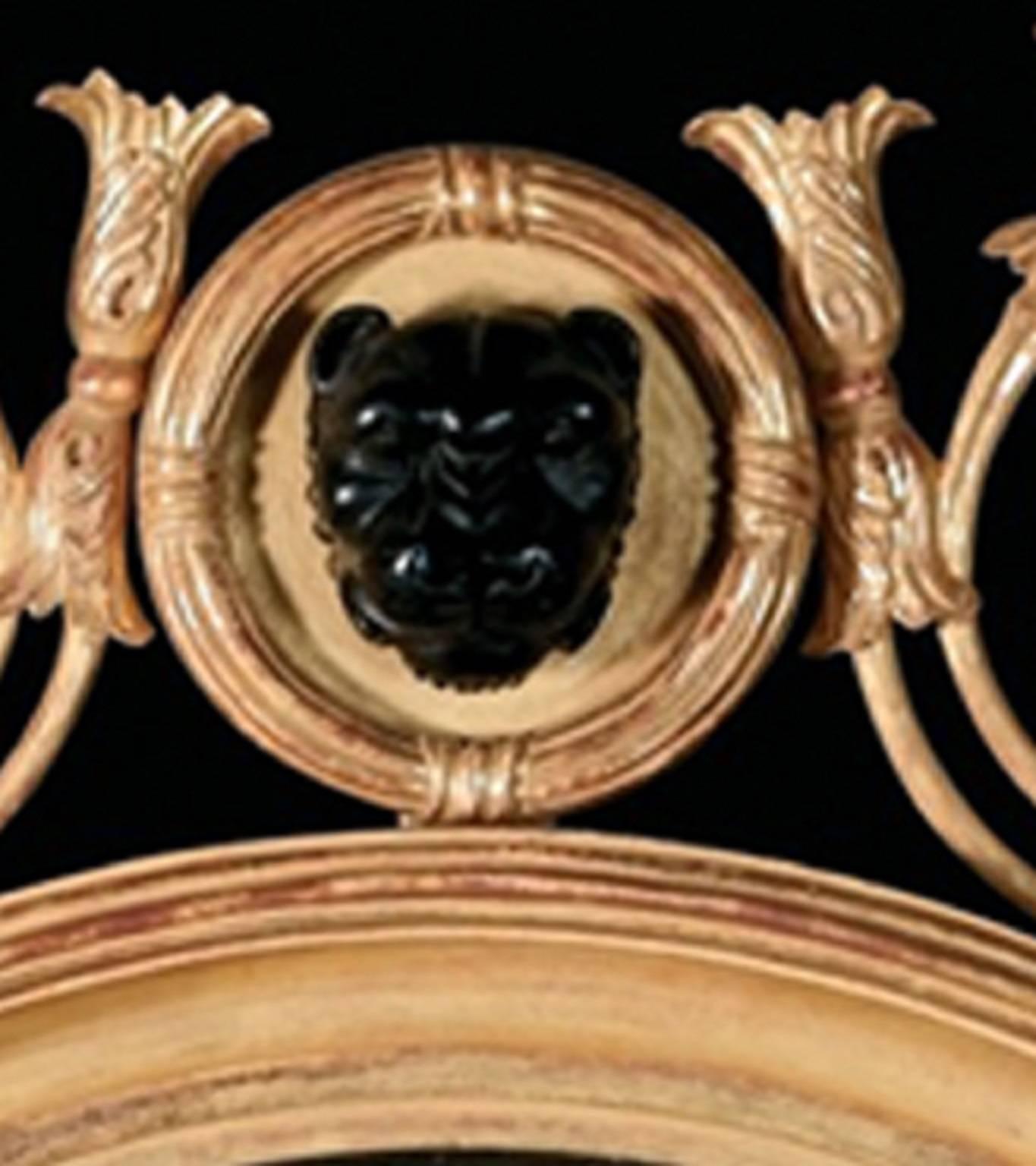This convex looking round glass mirror displays a wealth of Regency decoration. The carved giltwood, compound moulded outer frame has a reeded ebonized inner slip frame and a cresting centered by an ebonized leopard mask within a ribbon wrapped