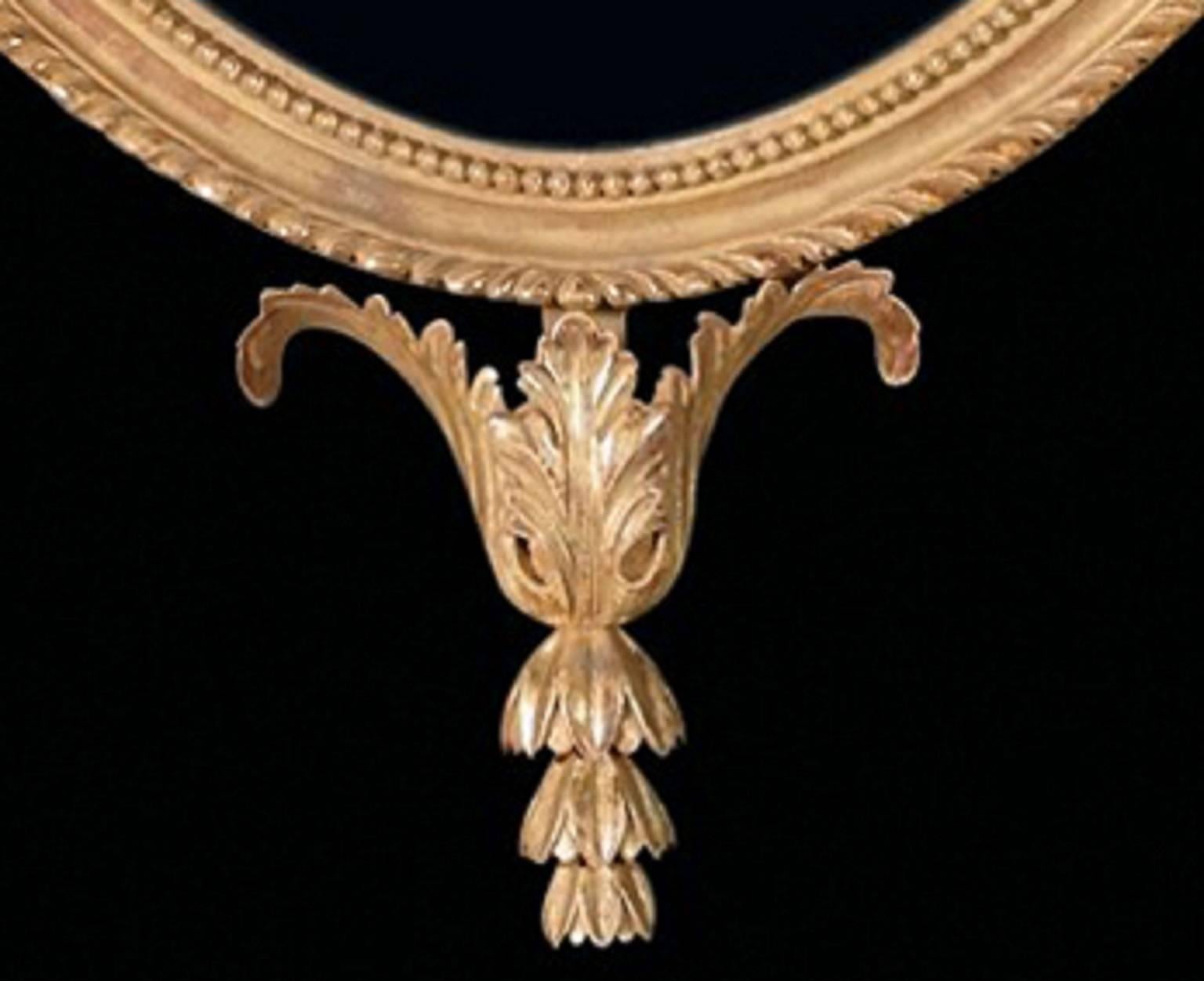 This oval carved and giltwood frame mirror, in the George III style of the late 18th century, is enriched by the acanthus leaf cresting supporting a tapered and fluted bracket, headed by an anthemion spray. 

The moulded frame is decorated with