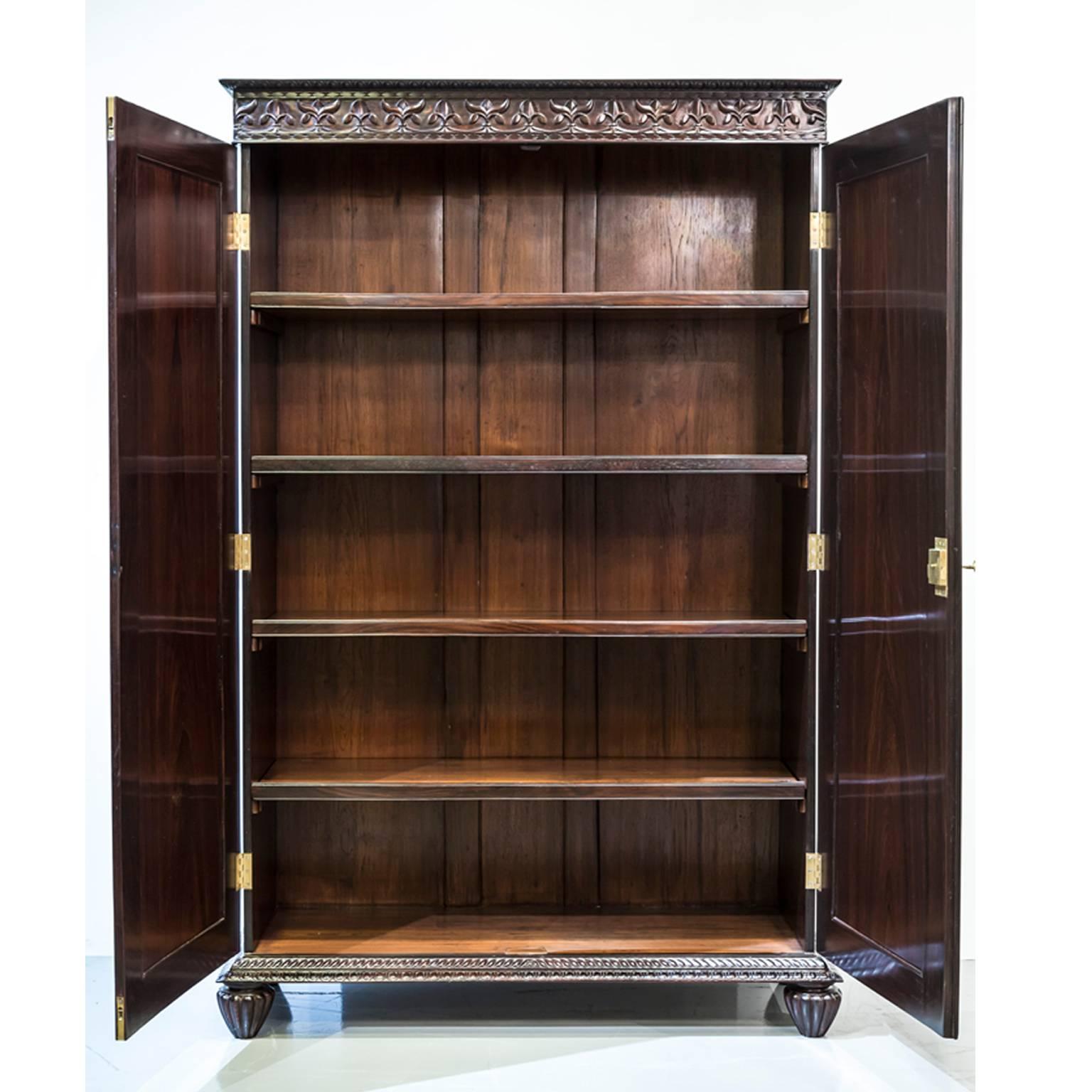 A British colonial rosewood cupboard with gadrooning on the edge of the flat top and Fleur-de-Lys carving on the frieze. 
The floating panels of the doors are carved with a large oval sunburst design, centred with an oval disk and leaf