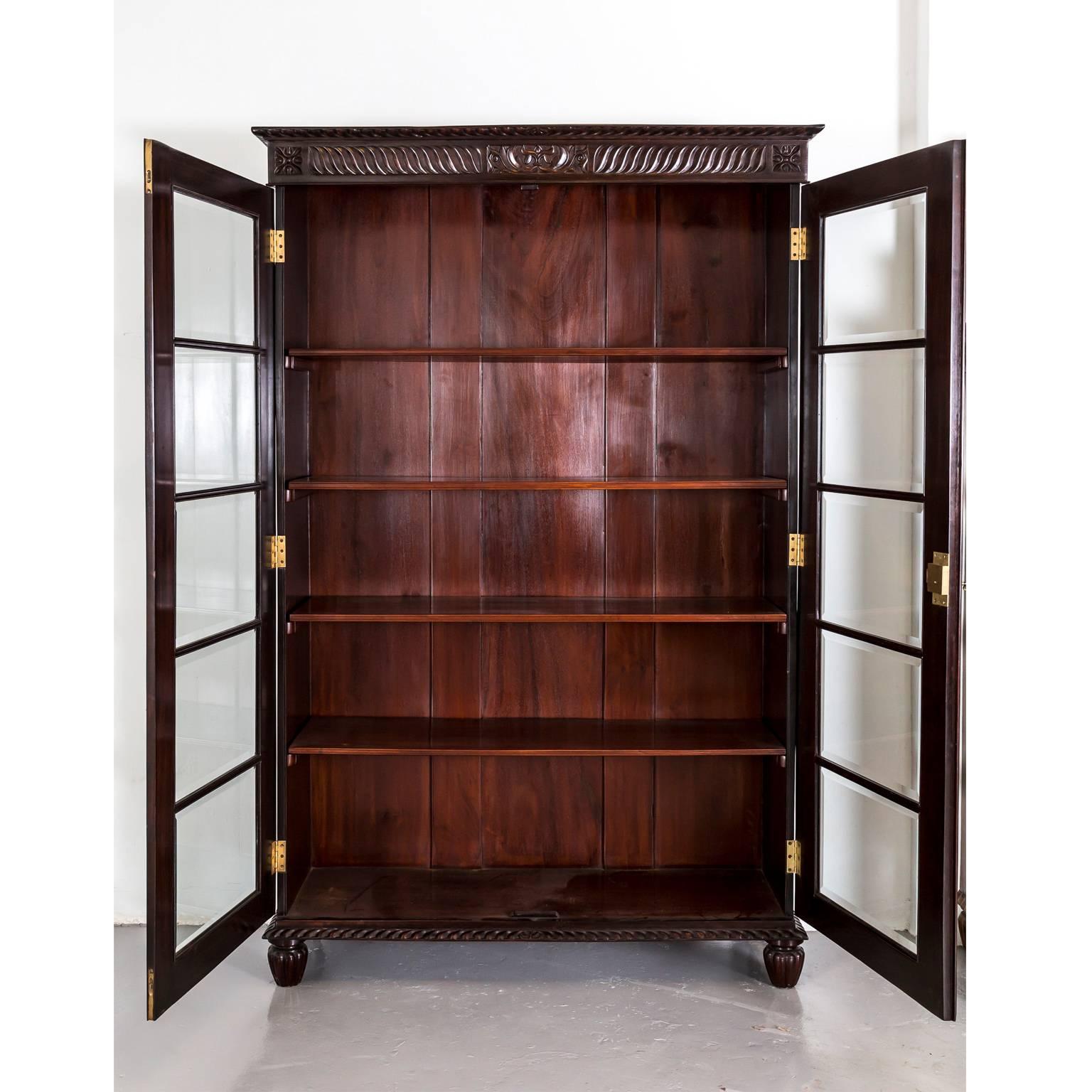 A British Colonial library bookcase in rosewood with a flat top, egg and dart carving on the edge and wave carving on the frieze centred by a floral block on the frieze. 
The frames of the double doors are carved with elongated reserves and each