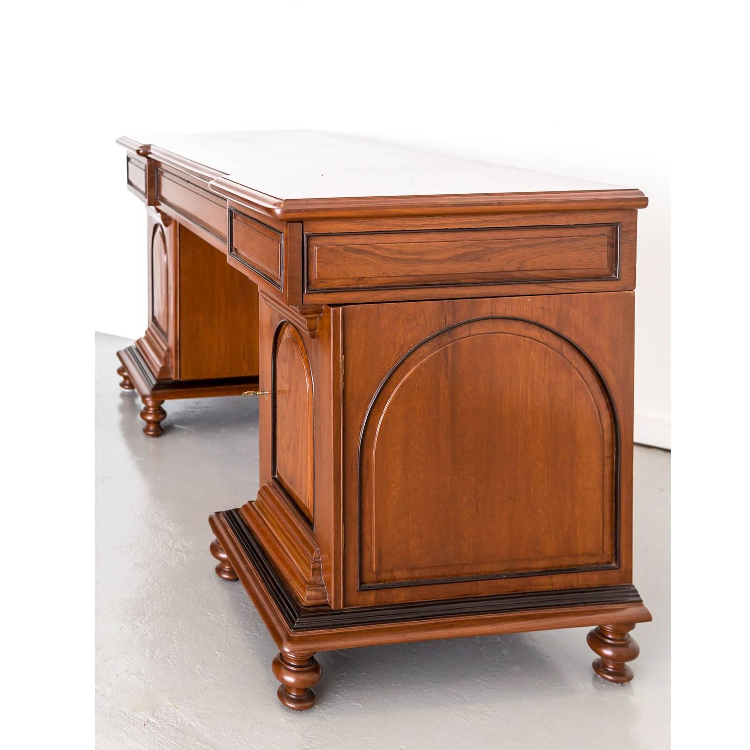 A British Colonial pedestal desk in teak wood with nicely contrasting rosewood details. 
The deep frieze features an inverted centre section with a large drawer flanked by two smaller ones. Both pedestals have a paneled door that opens to reveal