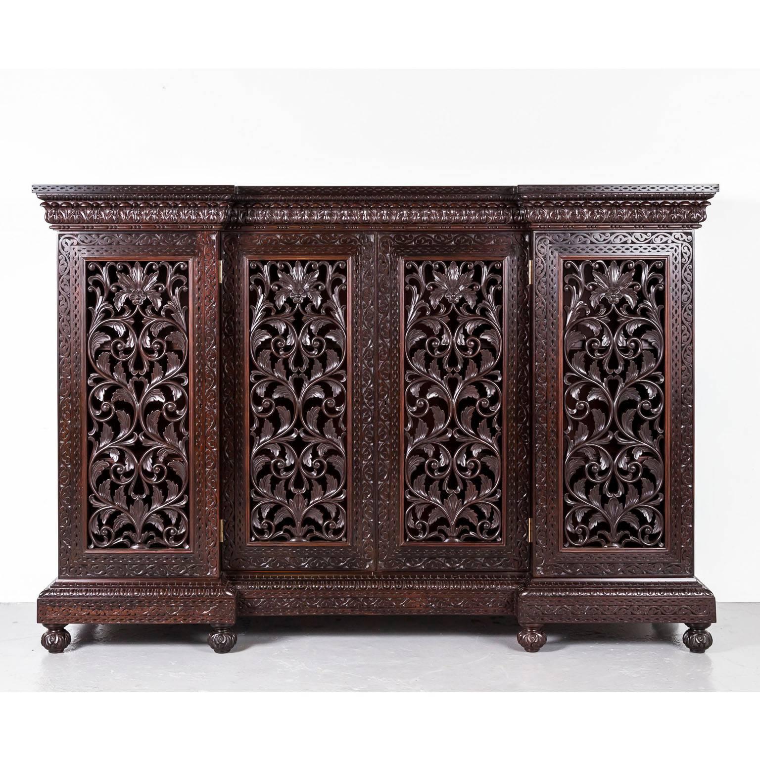 A beautifully carved and pierced British Colonial breakfront cabinet in rosewood. The overhanging top is made of one piece of rosewood and carved with two borders. The front is mounted with four hinged doors that are boldly carved and pierced with a
