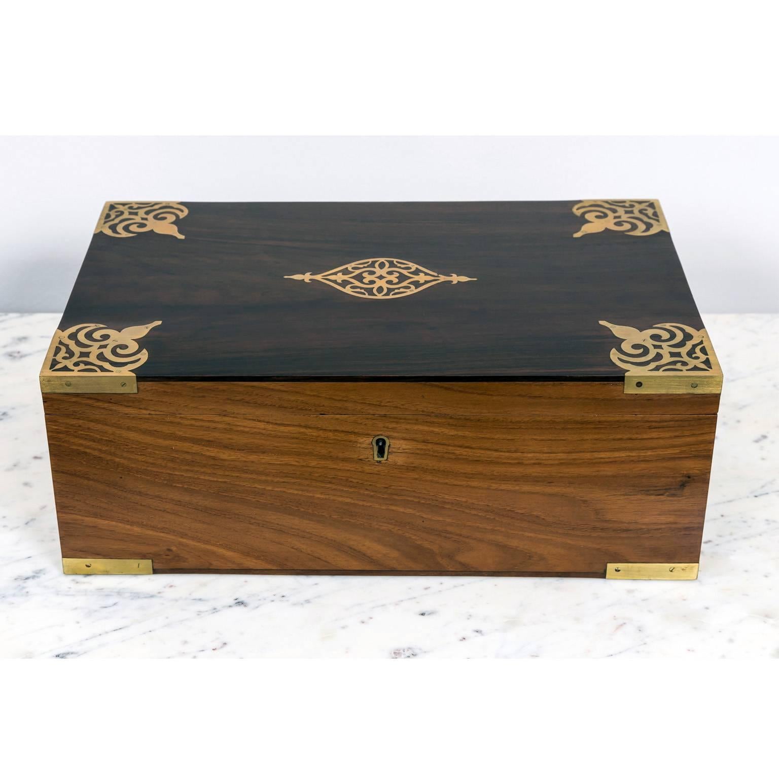 A beautiful British Colonial teakwood writing box with a brass lock, hinges and carrying handles. The lid of the box made of rosewood and decorated and reinforced with brass banding, corner mounts and a brass motif at center. 
The box opens to