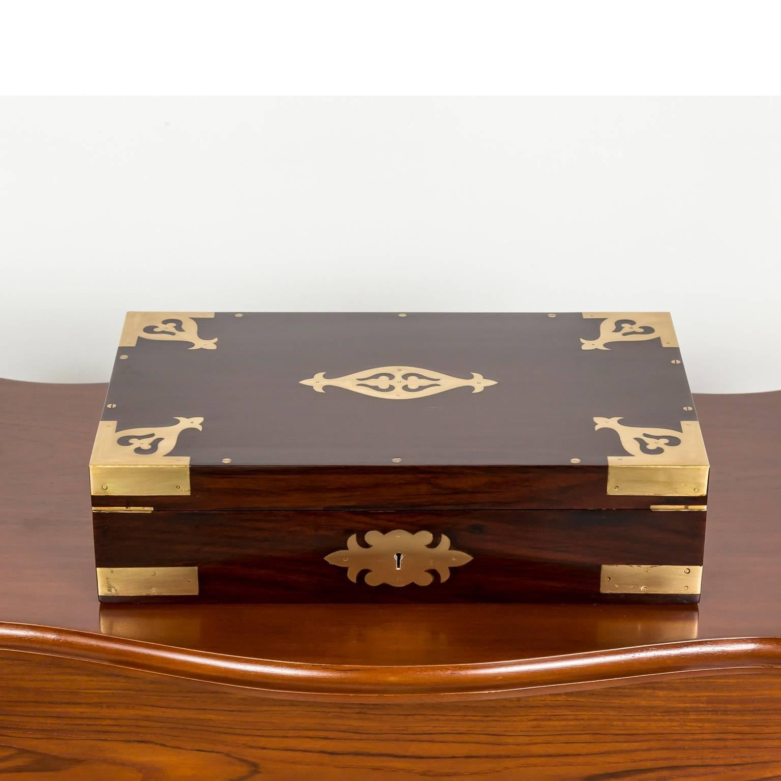 A British colonial rosewood writing box with beautiful brass Fleur–de- Lys decoration. Brass strapping to the edges, escutcheon plate and brass recessed lift handles on the sides. 
The box opens to reveal several small compartments. A large hinged