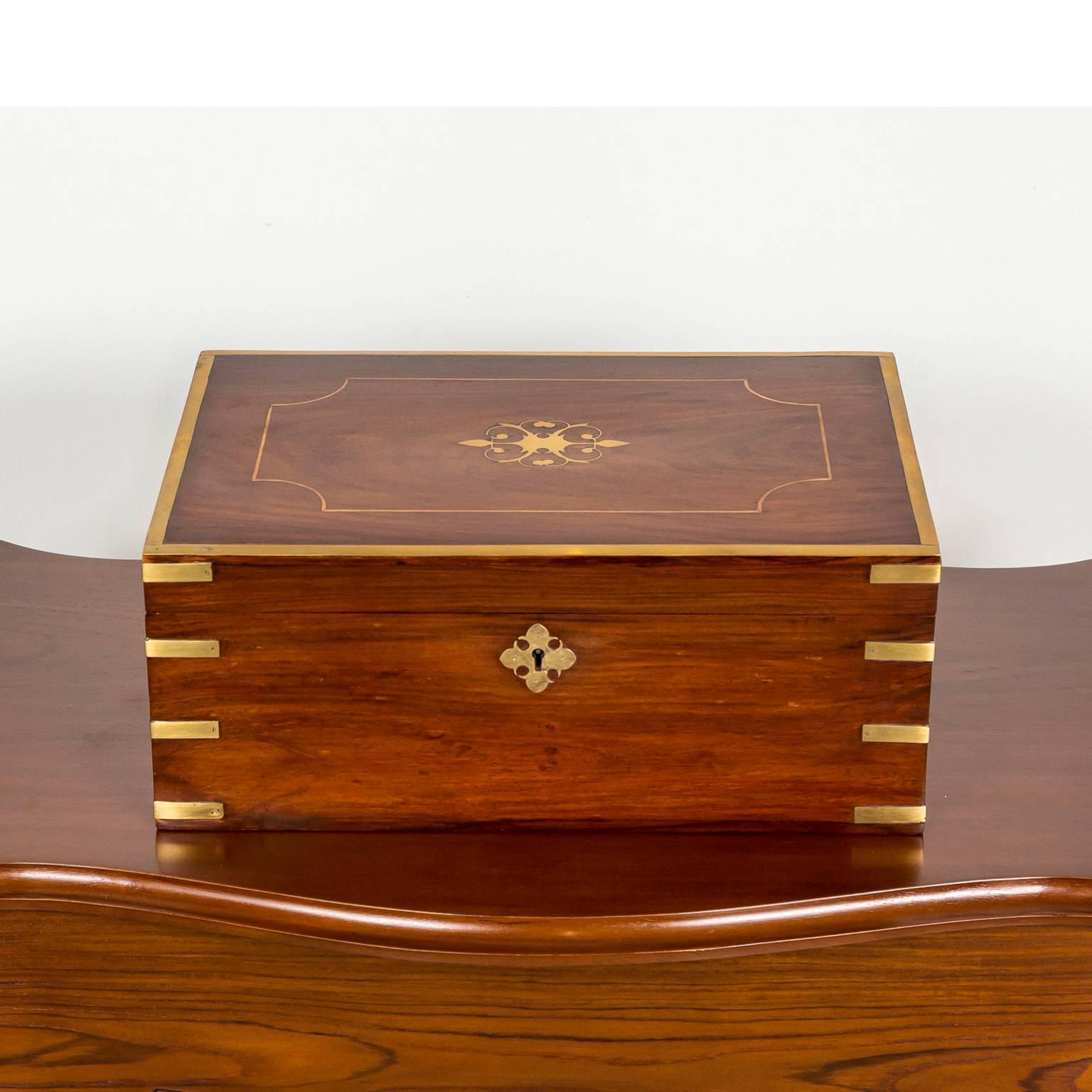 A beautiful British colonial rosewood dressing box with a brass ornament on the lid. The exterior reinforced with brass banding on the edges, both for decoration and protection. 
The box opens to a fitted interior with a mirror hinged on the