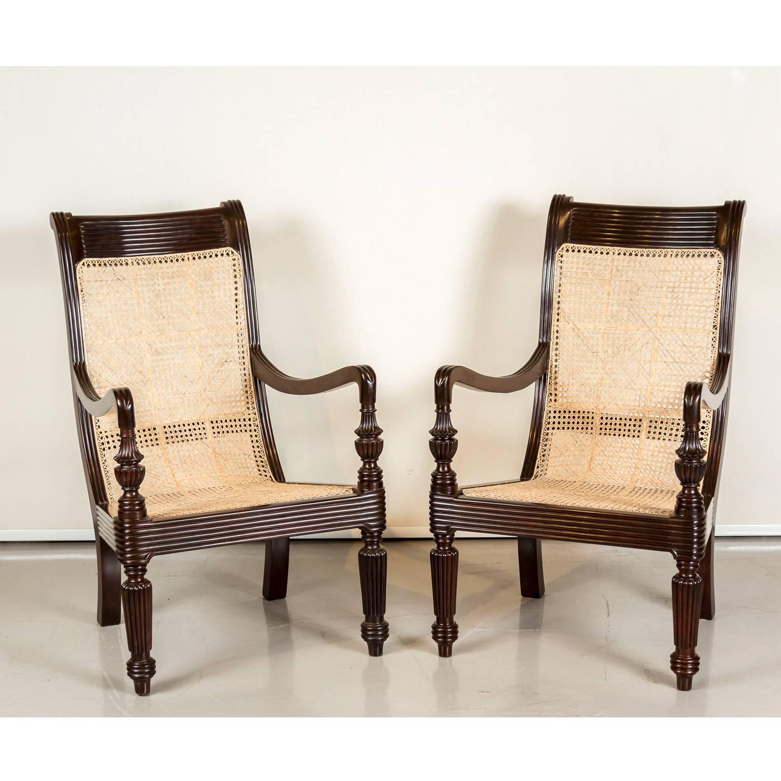 A pair of British colonial library chairs in rosewood with hand caned seat and back. The top rail and seat apron are reeded and the arms rest on flared tassel and ball supports. The front legs with multiple turnings and the rear legs slightly raked