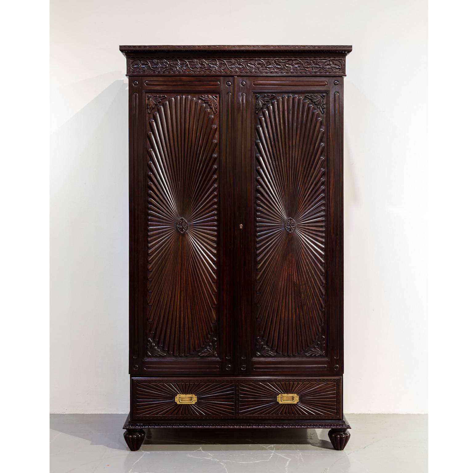A British Colonial rosewood cupboard with gadrooning on the edge of the flat top and intertwined floral carving on the frieze.
The floating panels of the doors are carved with a large oval sunburst design, centred with an oval disk and leaf