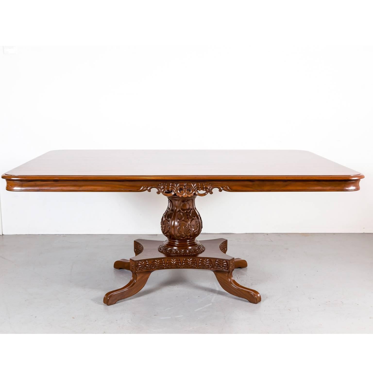 A rectangular British Colonial dining table made of teak wood. The top is figured with an edge above an apron with a nice decorative carving on the long sides.
It sits on a cylindrical turned pedestal with a beautifully carved baluster support that