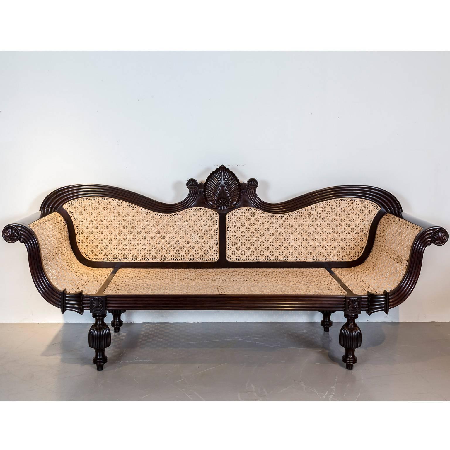 A magnificent British colonial sofa in rosewood with an arched serpentine crest rail centered by a floral carving. The reeded frame has scrolled arms that display finely carved floral roundels at the ends; a motif which is repeated above the legs.