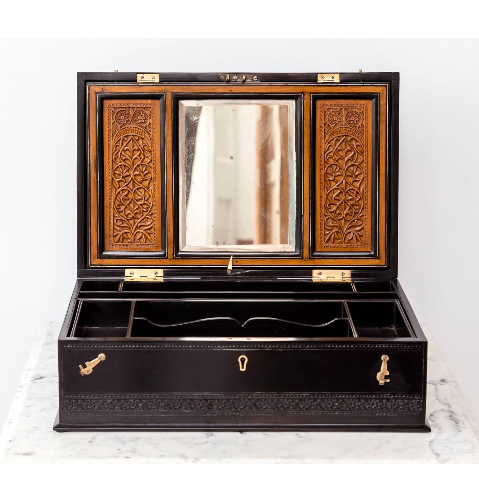 A beautiful British Colonial dressing box in ebony. On the top and sides the box has bands of foliage Moghul style carving. It opens to reveal an interior with a mirror flanked by beautifully carved foliage in sandalwood, several small compartments
