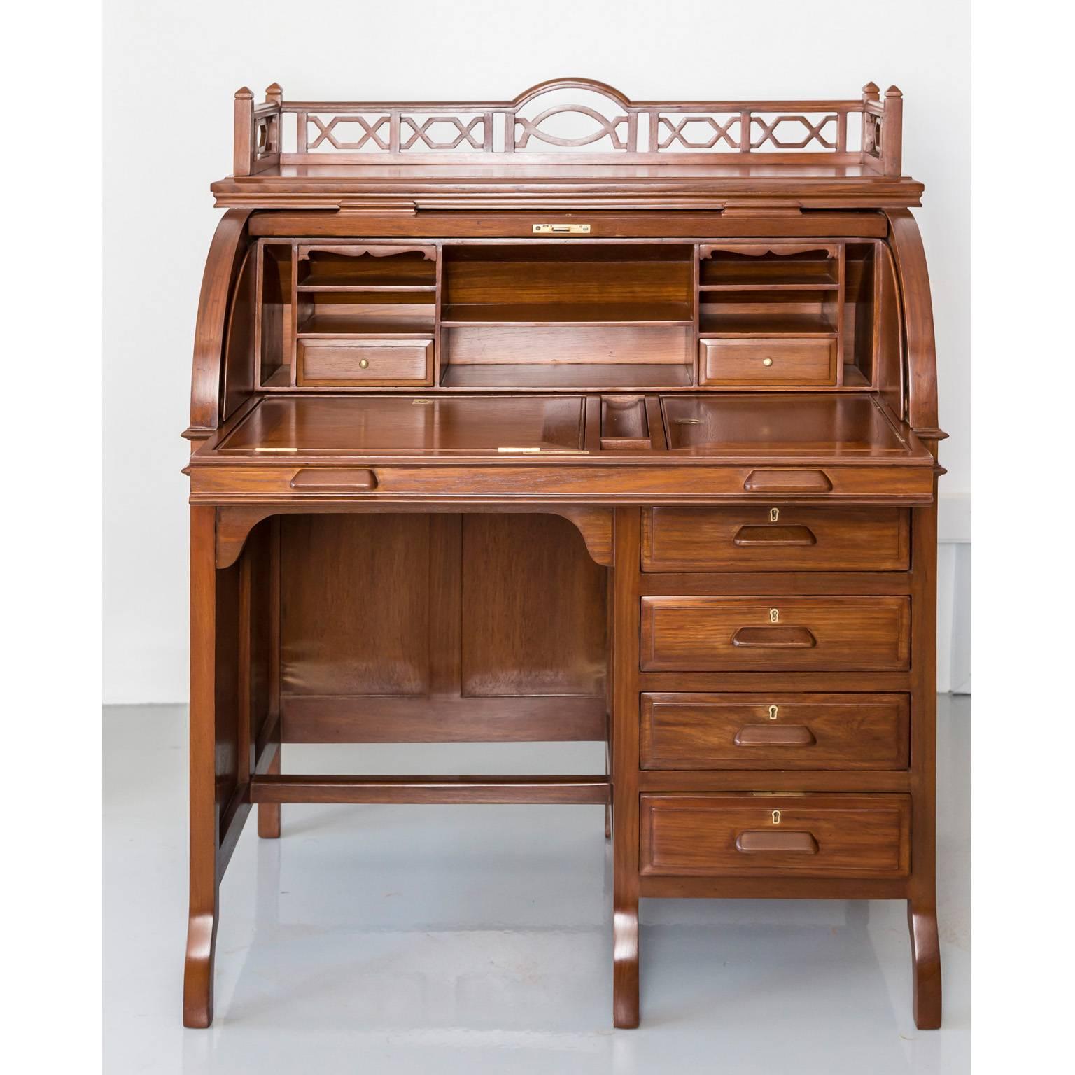 A British Colonial teak wood cylinder desk with a shaped back above a rectangular moulded top and a cylinder fall front. The top opens to reveal a fitted interior which consists of several pigeon holes and two drawers with brass knobs. In addition