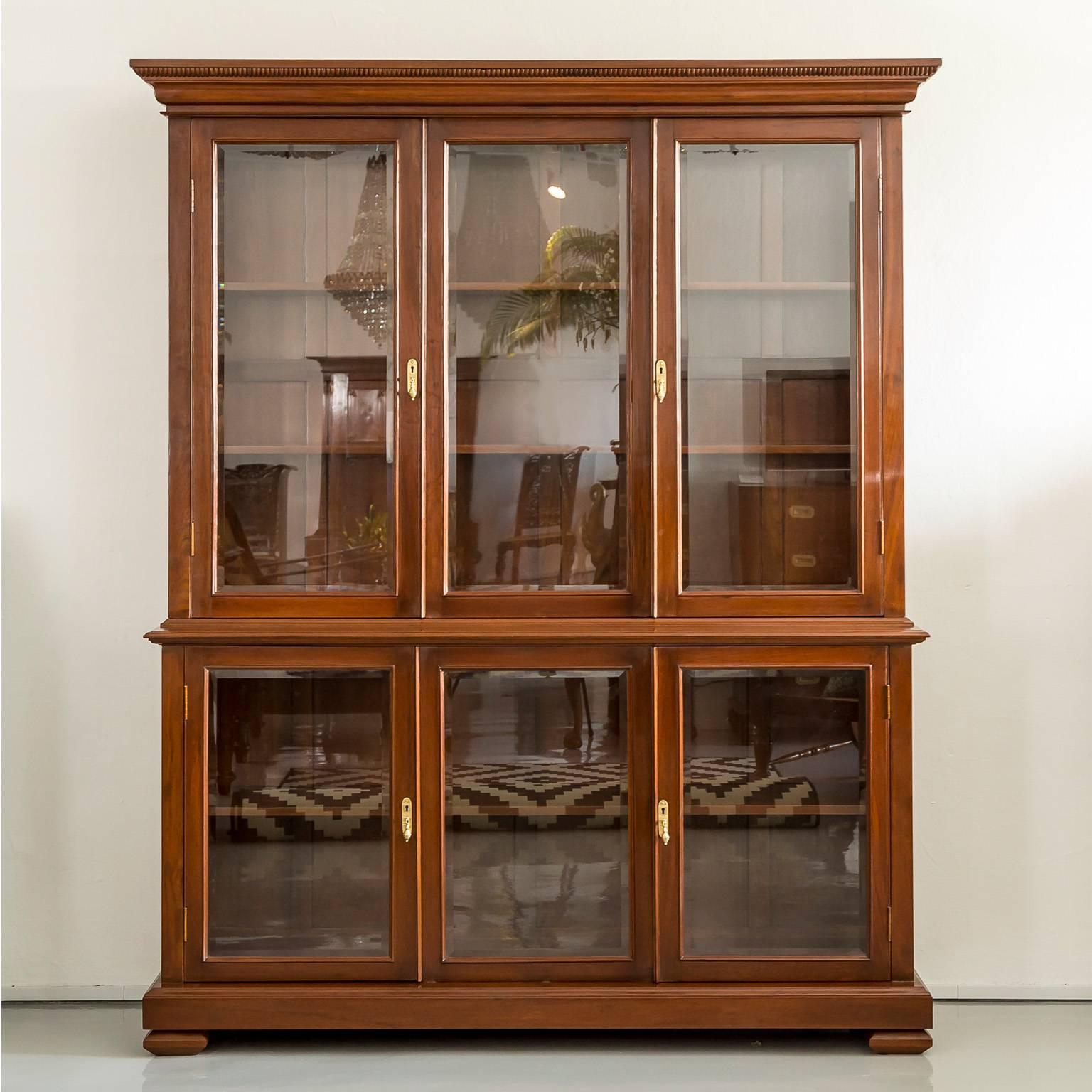 A British Colonial teak wood step back cabinet in two parts with a moulded cornice above a deep frieze. The upper section with a large glass panel flanked by a pair of glass doors, opening to two shelves. The deeper lower section also with a large