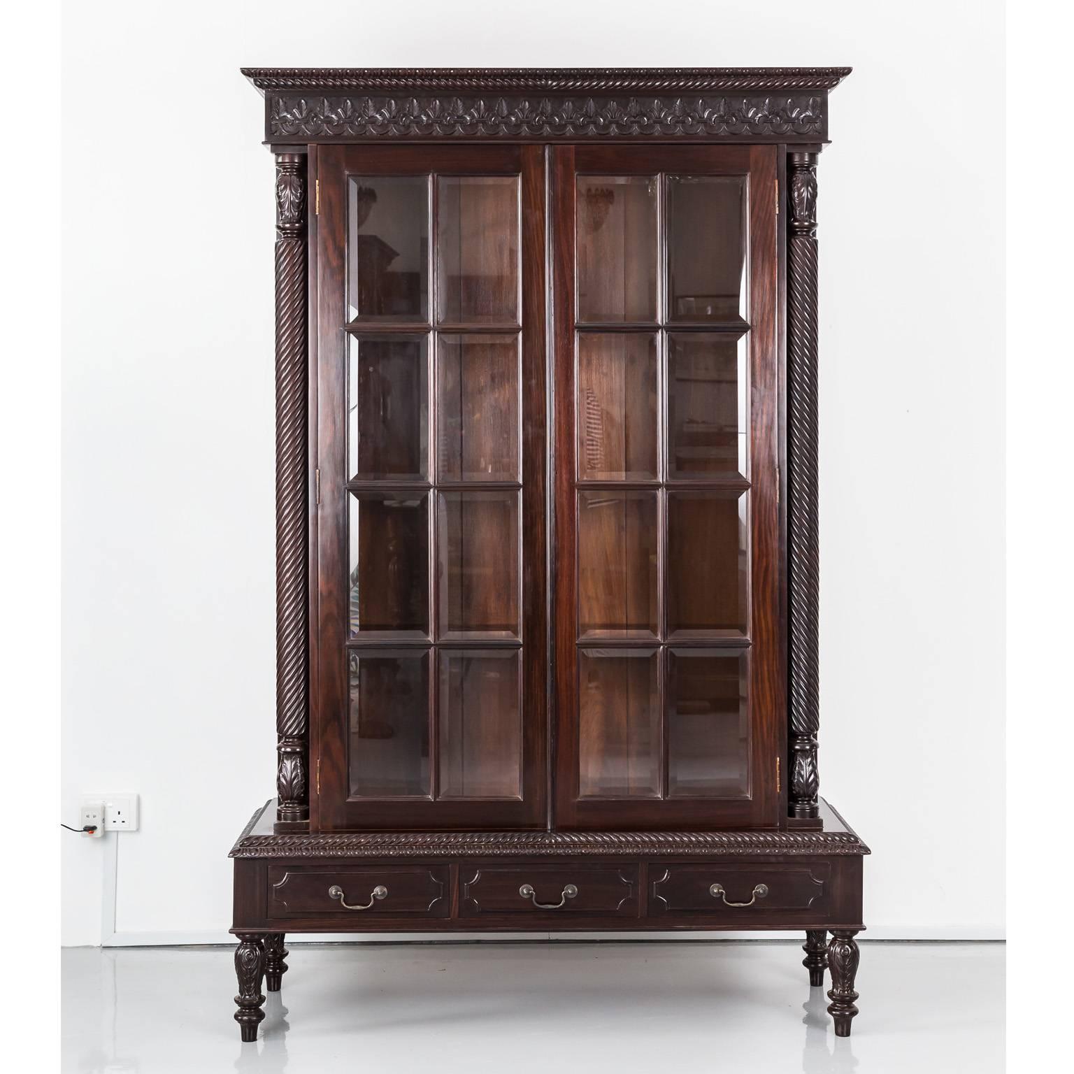 A beautiful British Colonial cabinet in rosewood with a deep moulded and carved cornice. The frieze is relief carved in a floral pattern. Set back from the base, the cabinet has double doors with eight bevelled glass panels flanked by two spirally