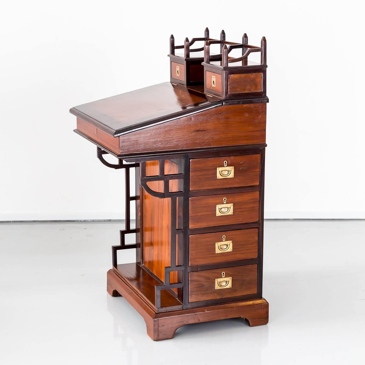 A British Colonial Davenport desk in teak wood with nice contrasting details. It has the original wooden gallery above a writing slope that opens to reveal storage space. 
On the rectangular top a gallery with two small drawers with brass ring