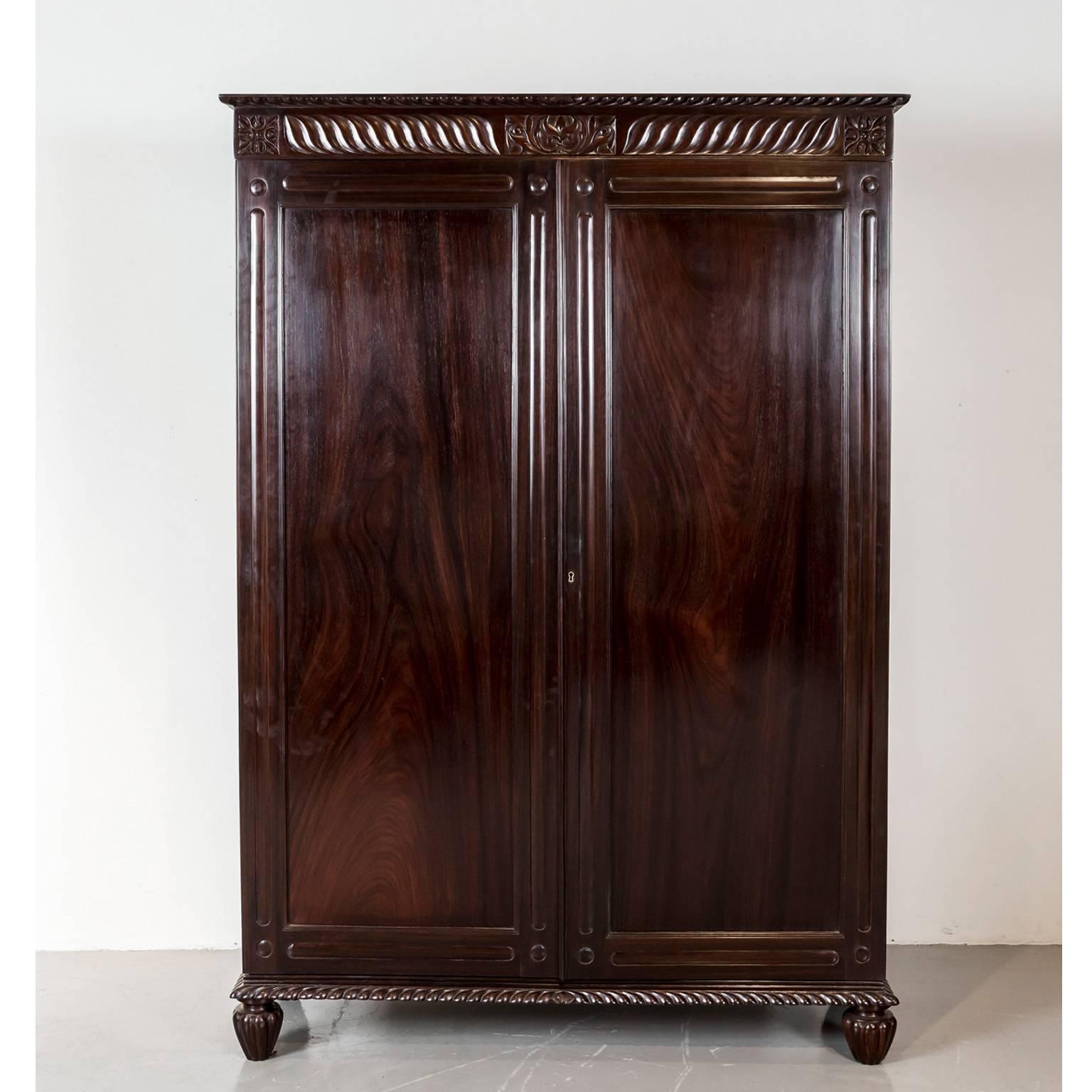 A British colonial cupboard in rosewood with a flat top, egg and dart carving on the edge and wave carving on the frieze centered by a floral block. 
The frames of the double doors are carved with elongated reserves and circles in the corners, the