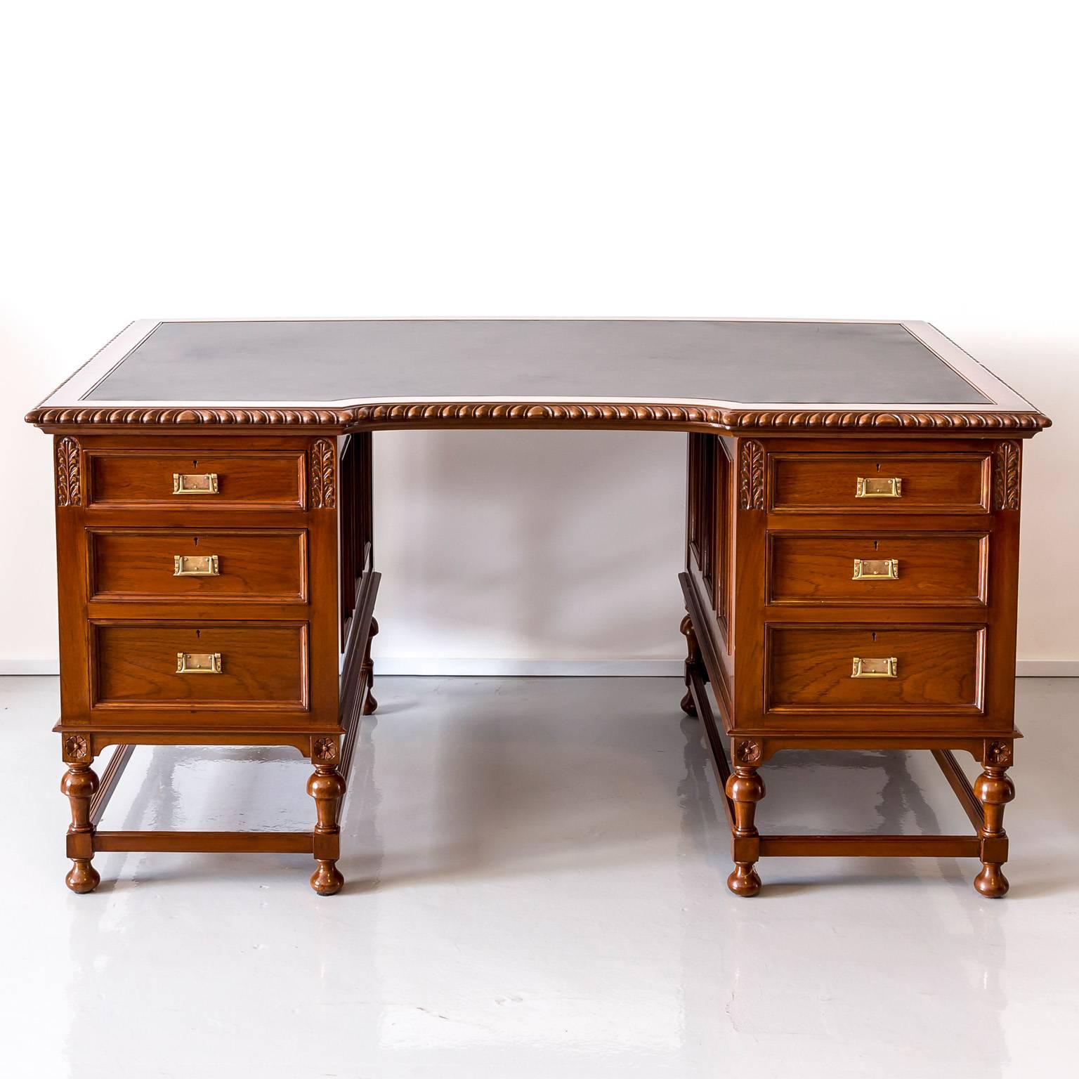 An elegant British Colonial pedestal desk in teak wood with a new inset black leather surface within an overhanging top panel. The top features an inverted centre section that is flanked by two pedestals. Each pedestal has three graduated drawers,