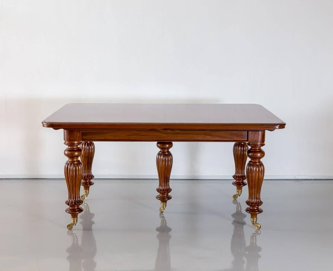 A British colonial teakwood extending dining table with rounded corners. The top, with a deep moulded edge, extends well beyond the base. 
The table has a telescopic under-frame that extends via a winding mechanism, which allows the table to be