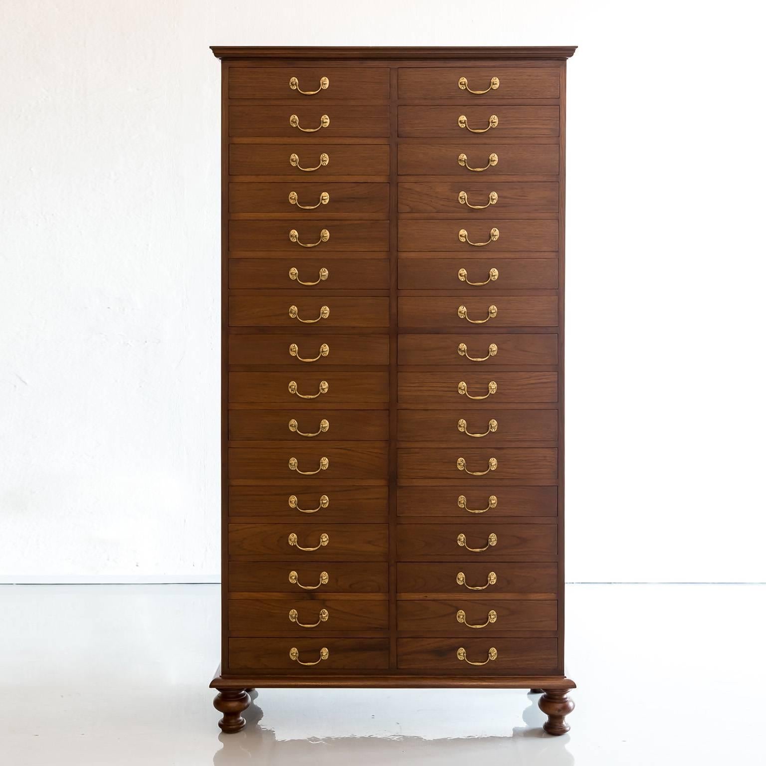 A British Colonial multi-drawer cabinet made of Burma teak wood. It features an overhanging moulded top above two rows of 16 drawers of equal size, all drawers with swan neck brass handles. The cabinet rests on a base that stands on four legs. Due