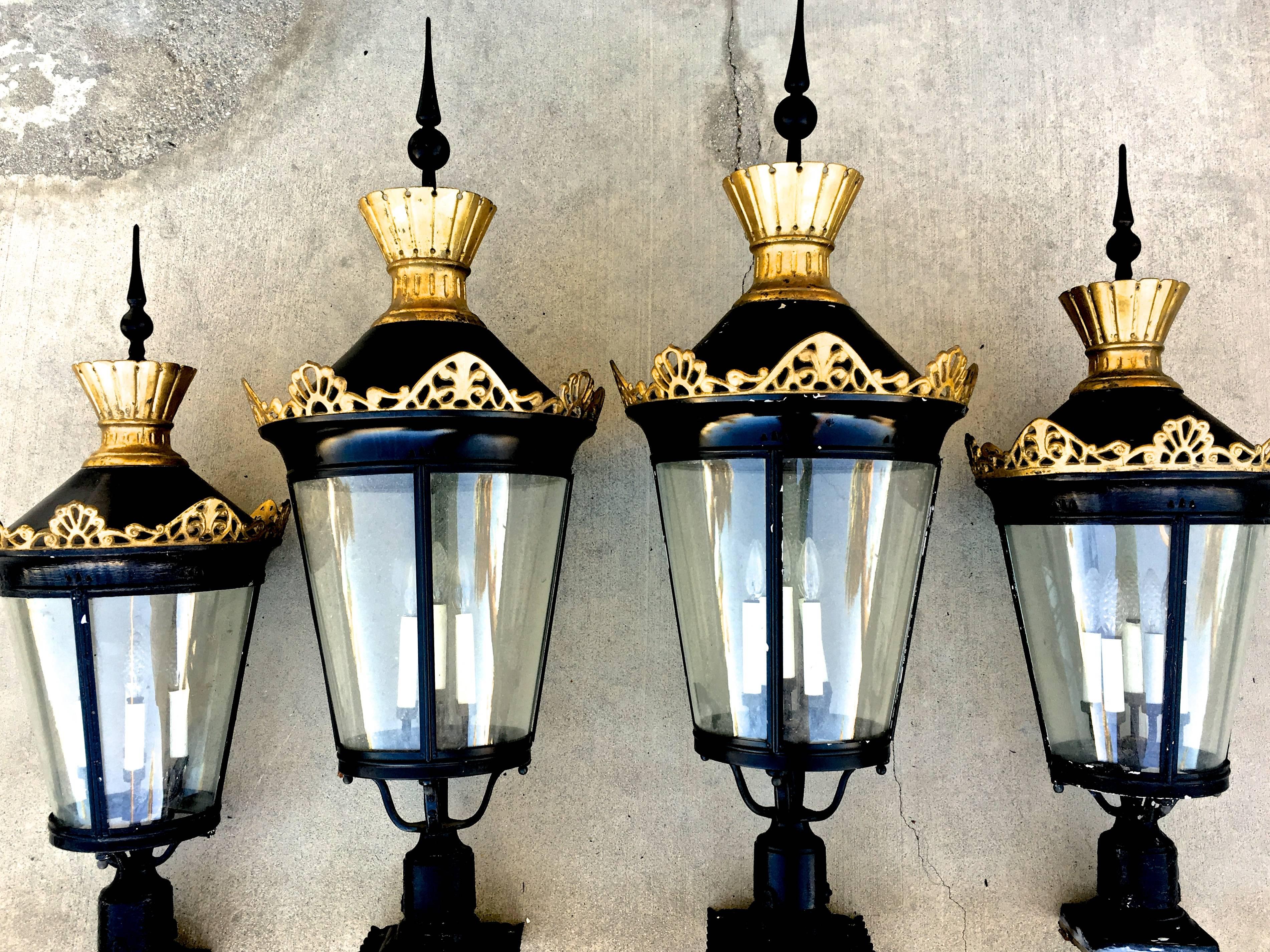 Dramatic scale and spire-like finials define this unique set of four Hollywood Regency post lanterns. Completely intact and operating, the antique fixtures are made of painted metal, glass, and feature gilded brass accents, each with a five-arm