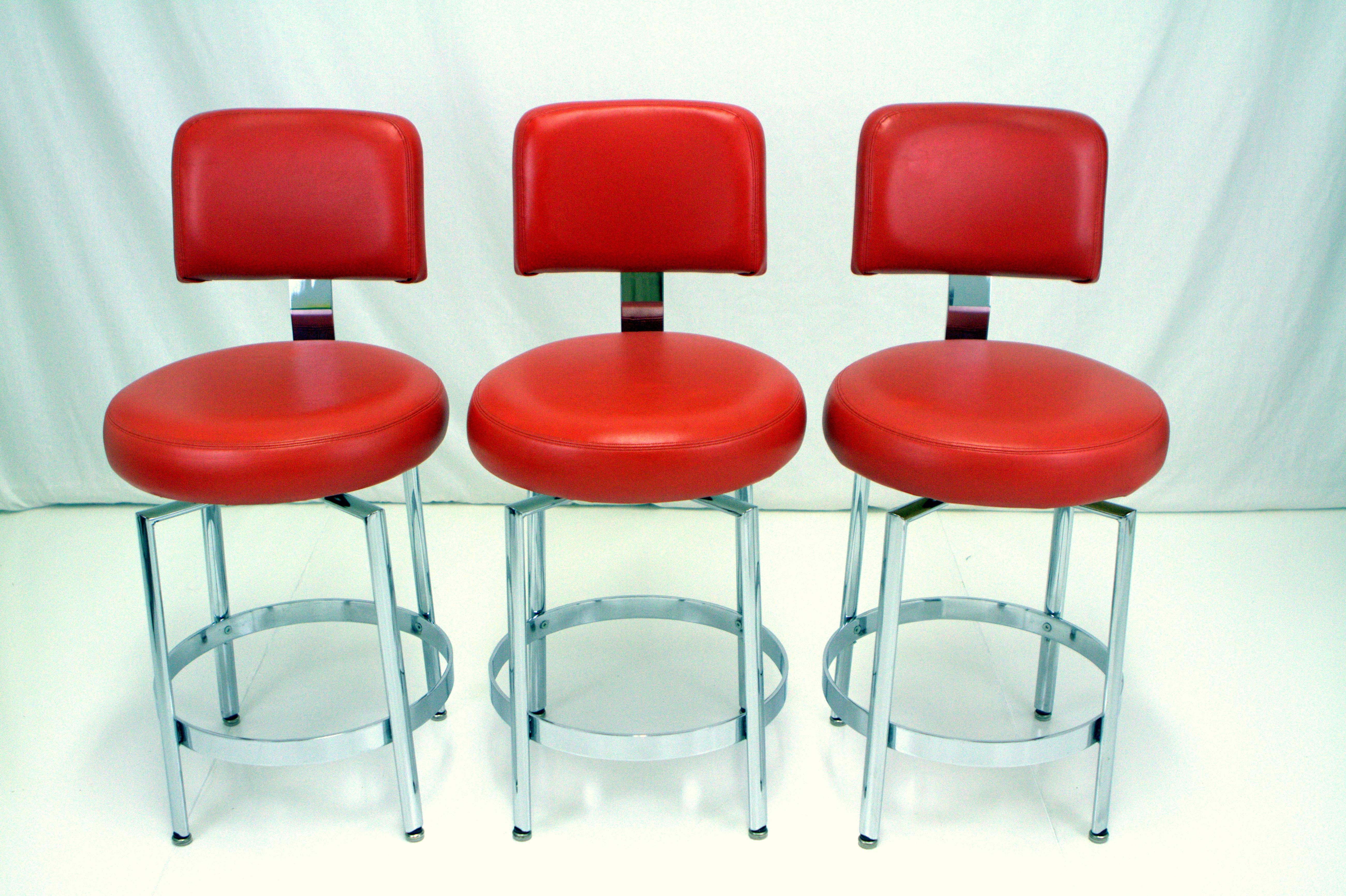 Modernist set of three chrome and orange vinyl counter height stools by Tri-Mark Tulip. Comfortably upholstered round seat with generous back support on chrome swivel base with round foot rest.  