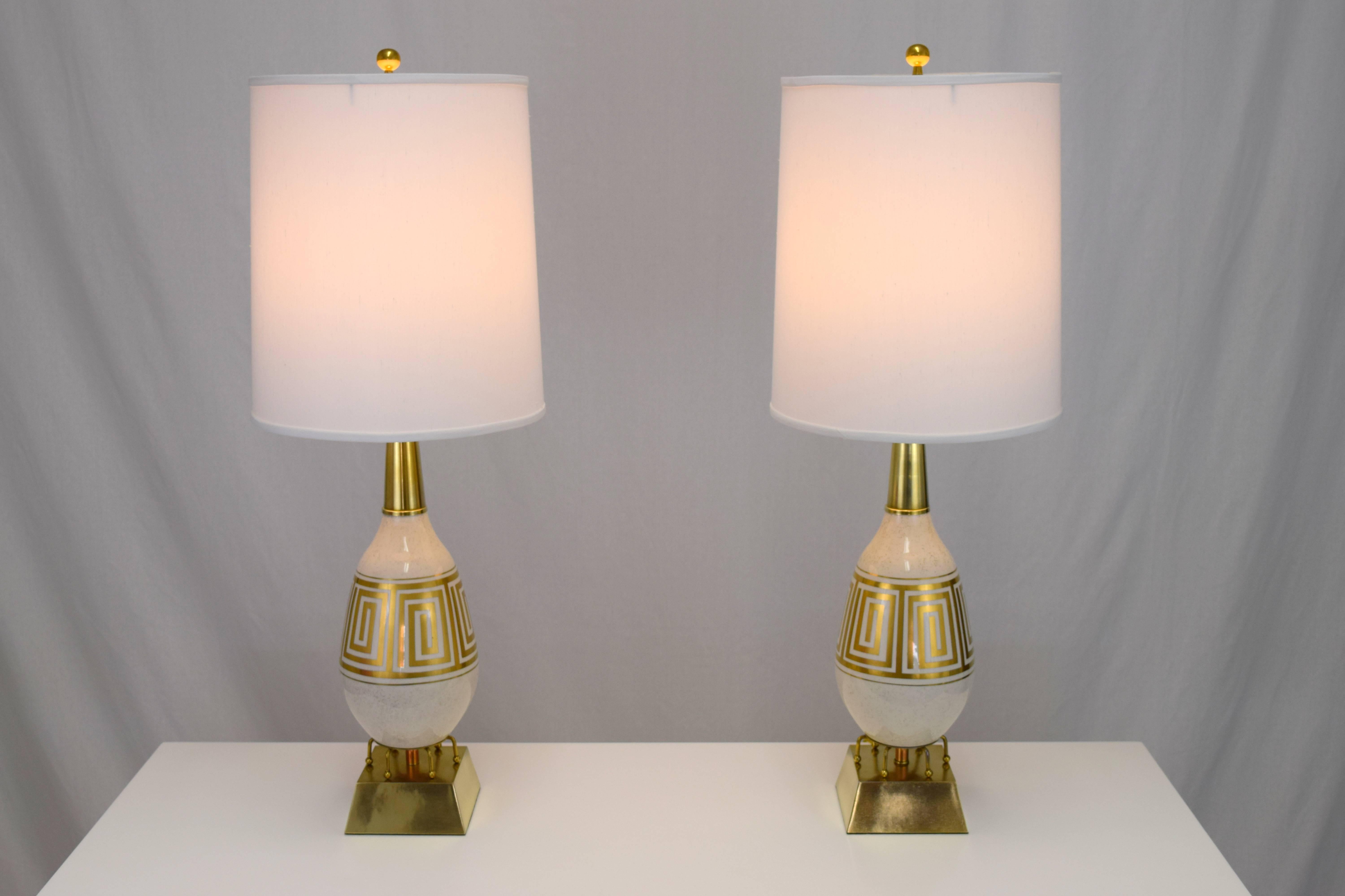 Amazing pair of ceramic and brass lamps with a Greek key motif. Egg shaped ceramic body in a cream and gold speckled glaze with gold leaf Greek key band. Six armed ball feet rocket ship like base resting on pyramidal block in brushed brass. Height