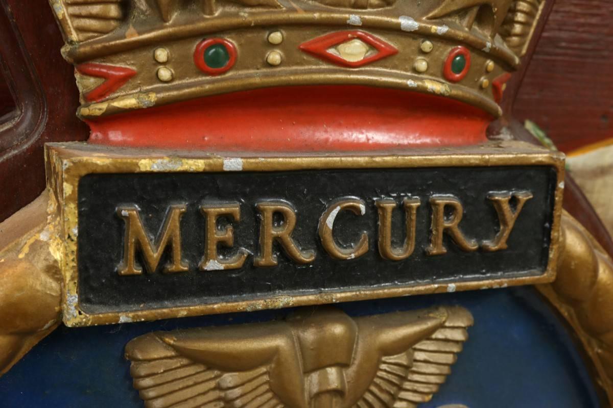 Ship's coat of arm badge from the HMS mercury. The metal badge is hand-painted, the arched top crown shows ships sterns. The crest shows two serpents climbing a safe and ship name mercury across badge. Mounted to hardwood backboard with routed edge.