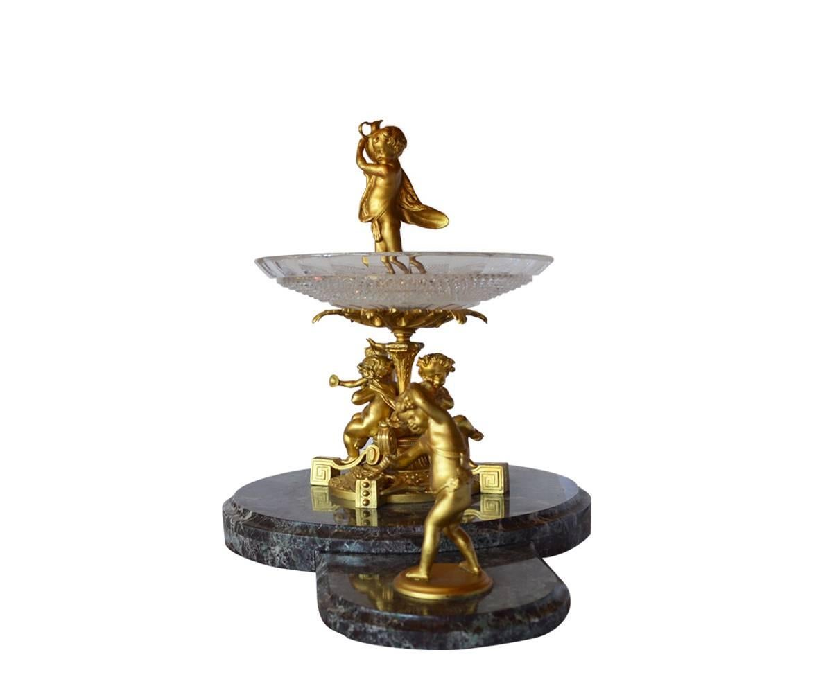 Five animated Putti adorn this centerpiece with two playing on the base which supports the cut glass bowl into which a third Putti is preparing to fill the bowl from his urn. Two more dance at the perimeter, one on each side. This piece is unusual