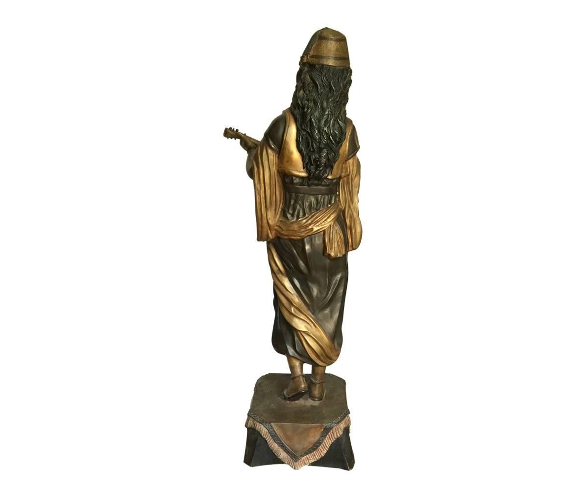 This is a bronze female with a flowing dress, scarf and vest playing music and standing on a decorated pedestal, circa 1930.